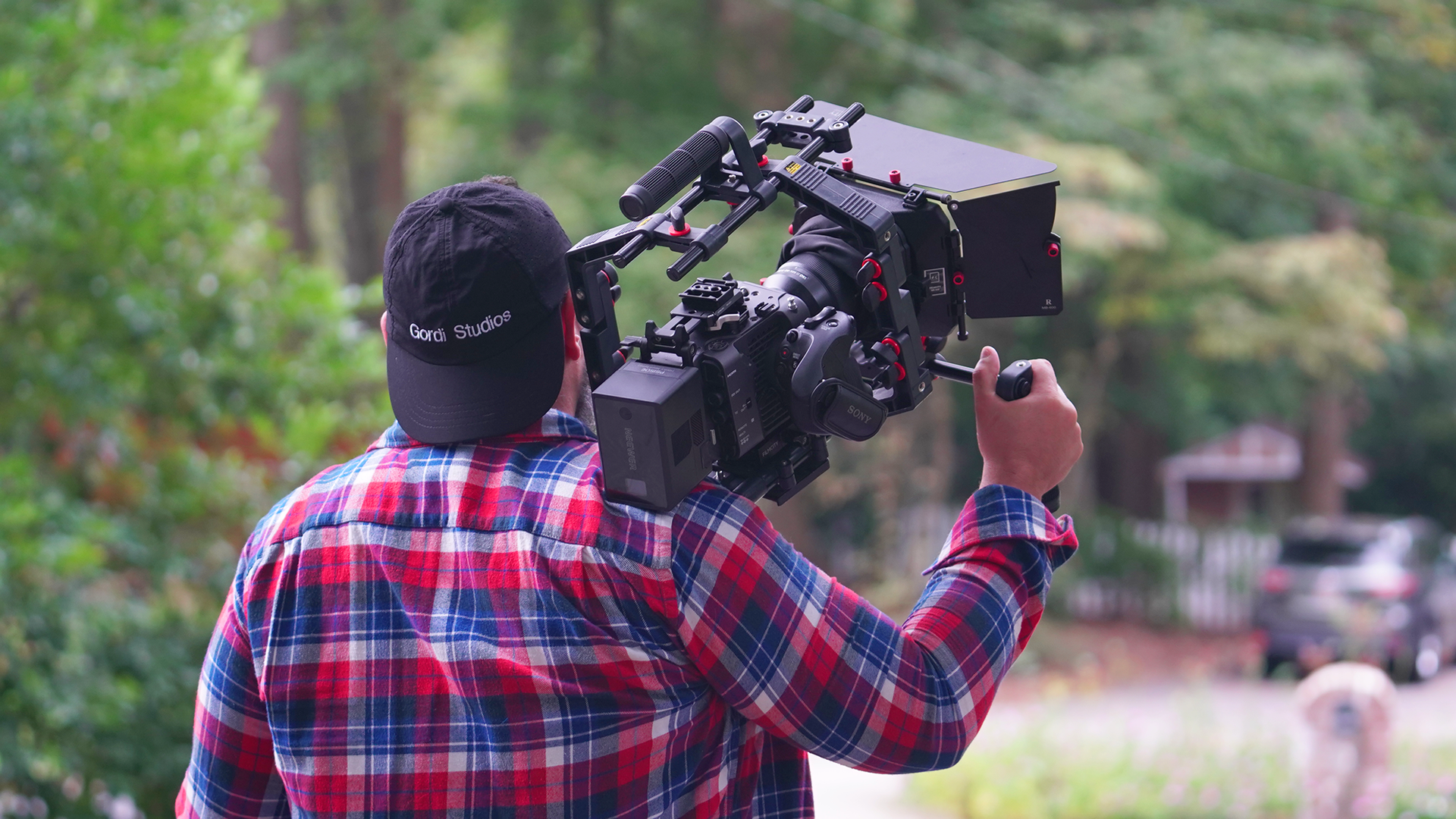 Cameraman in a red plaid shirt filming outdoors with a shoulder-mounted video camera