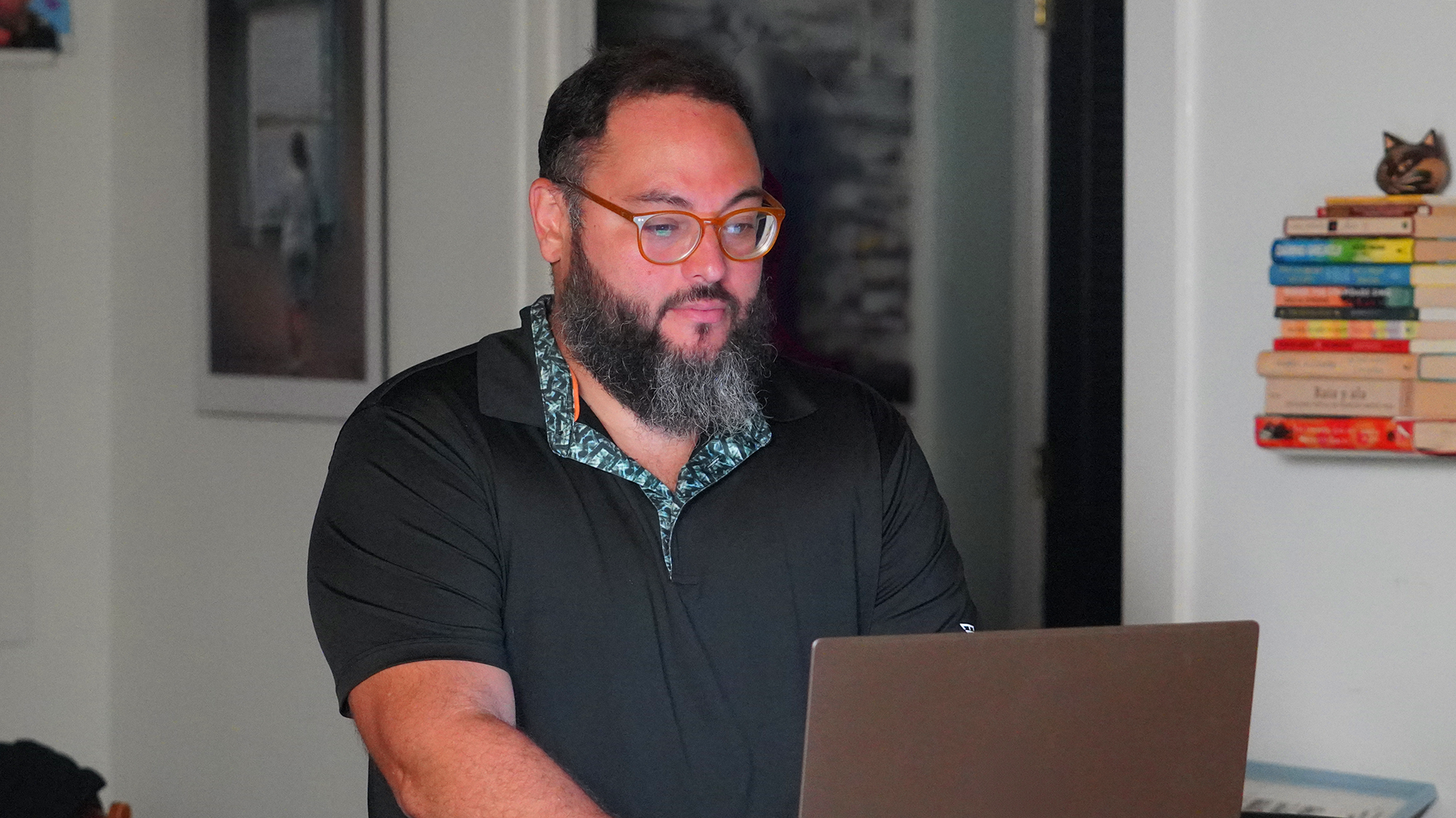 Person with glasses and a beard using a laptop indoors beside a stack of books.