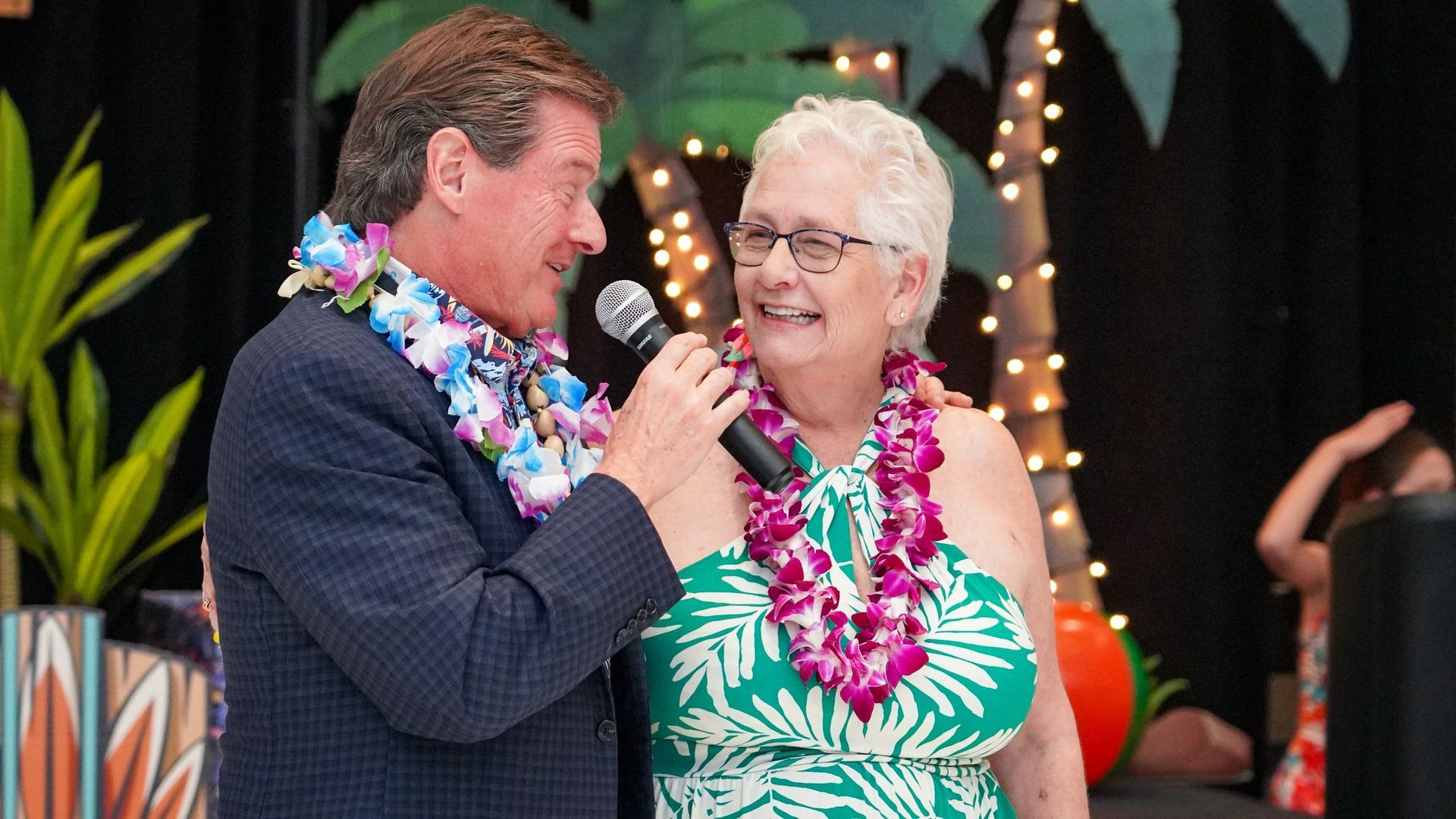 Man speaking into a microphone to an elderly woman wearing leis at a festive event