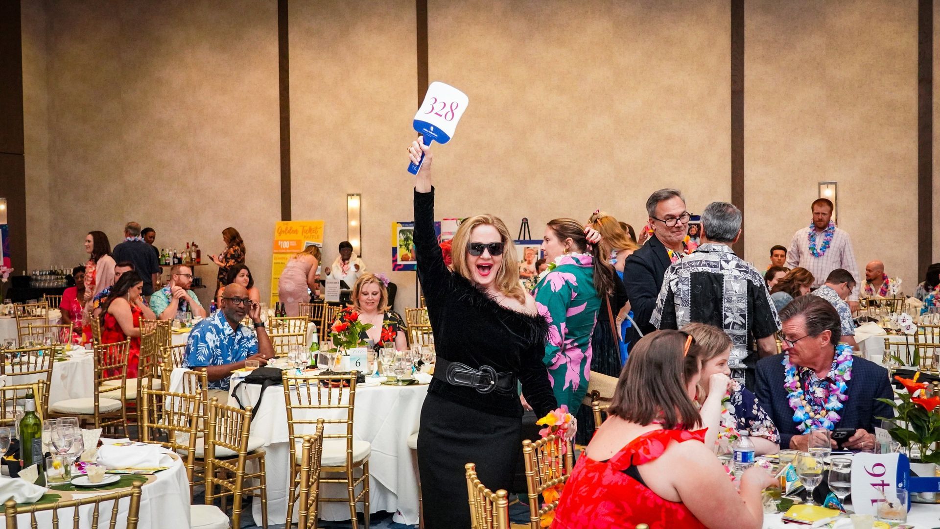 Woman in black raises a small award at a banquet table in a crowded event hall.