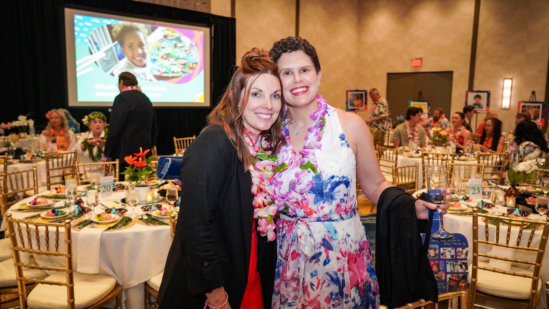 Two women smiling at a banquet table in a decorated event hall, with a presentation screen in the background