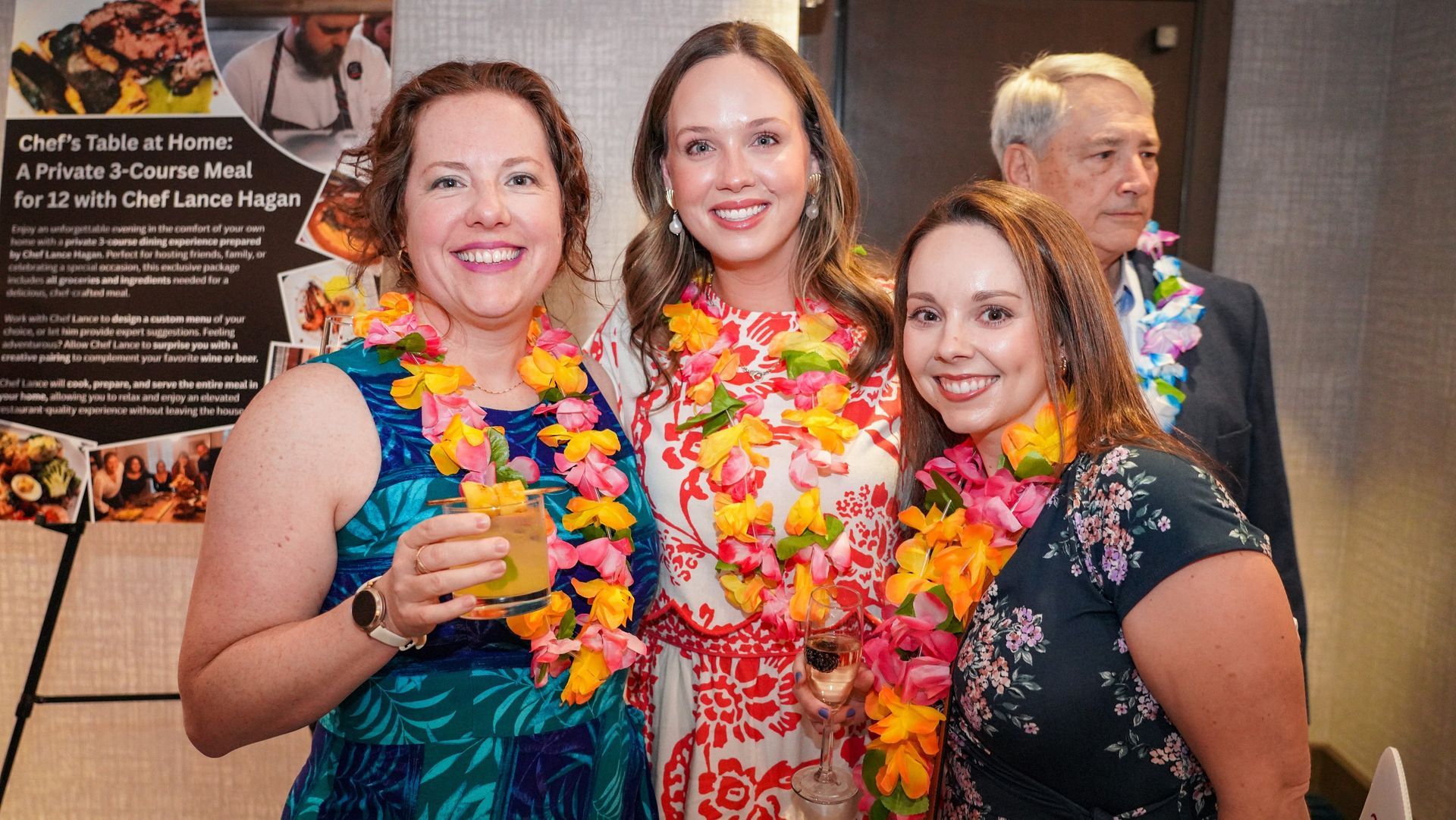 Three smiling people in floral leis pose at an indoor event, holding drinks and standing by a wall display.
