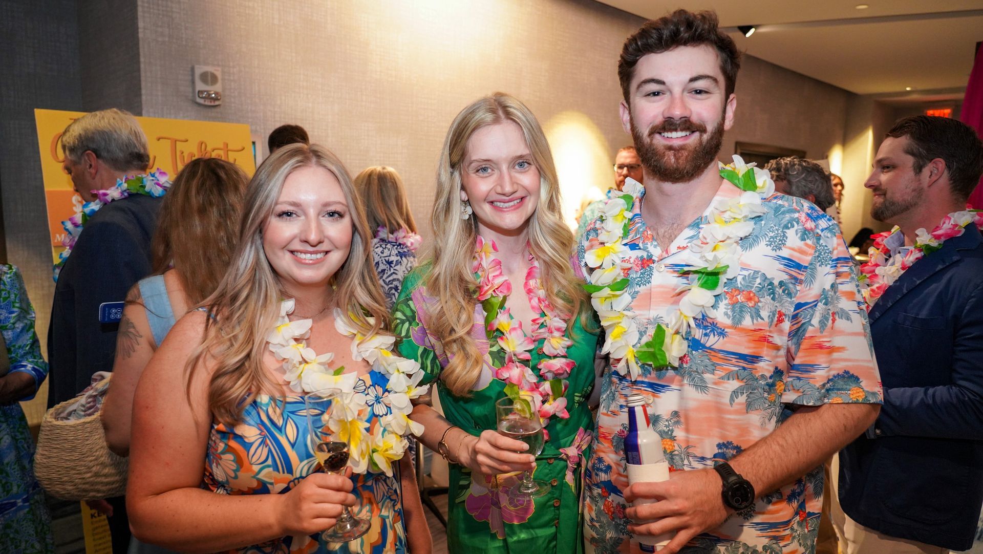Three smiling people in colorful leis posing at an indoor party.
