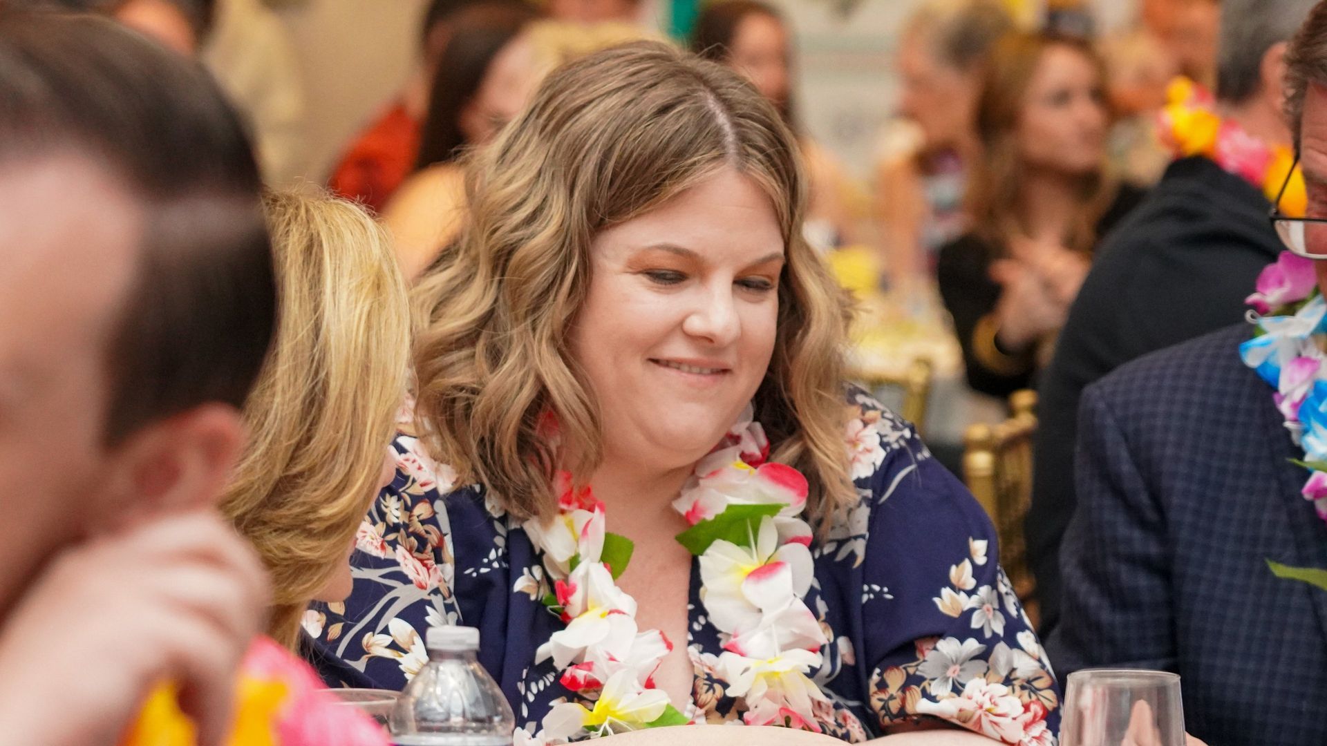 Woman smiling at a festive dinner table, wearing a colorful flower lei amid a crowd.