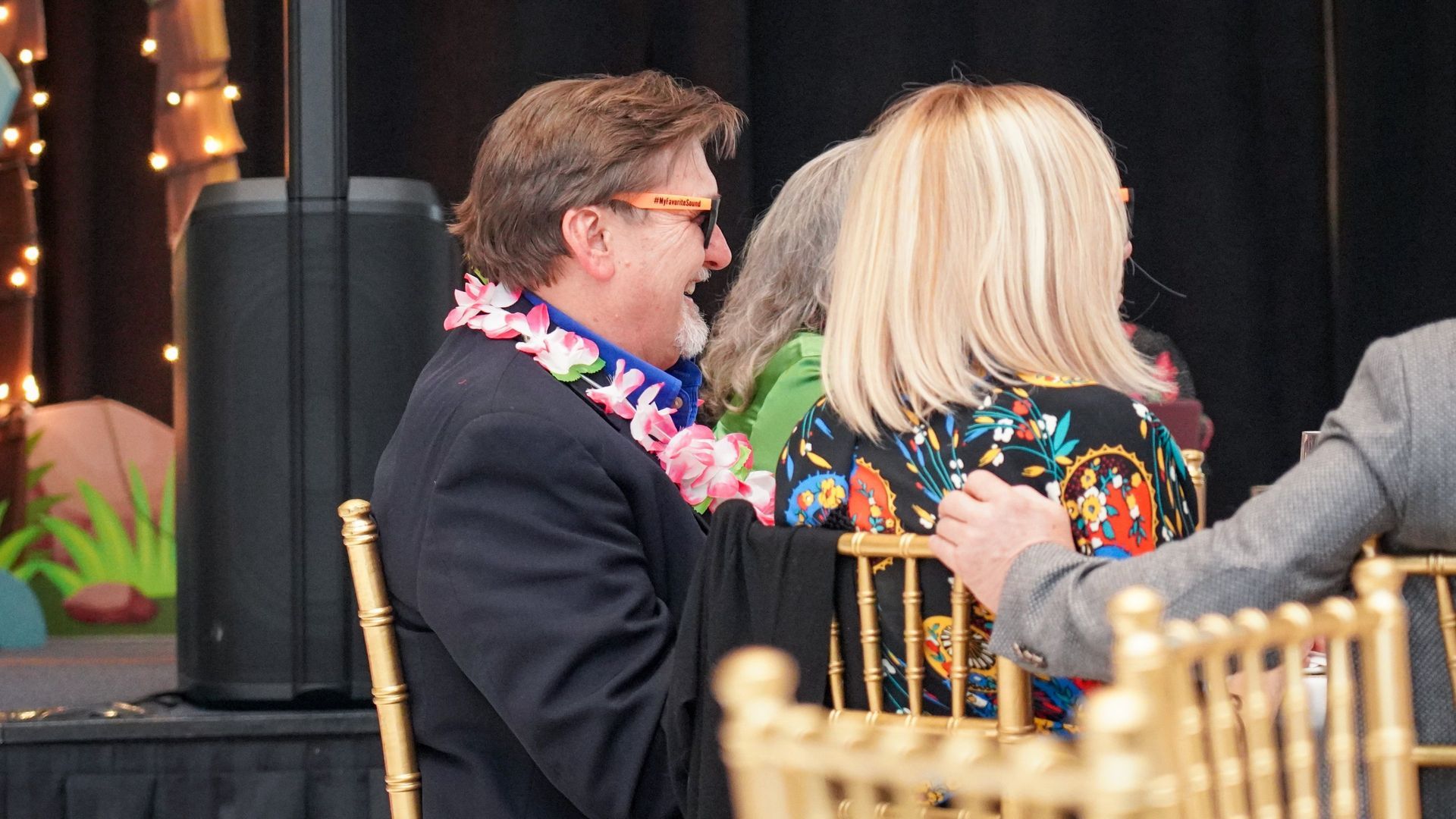 People seated at a formal event, one wearing a floral lei, with gold chairs and a dark stage backdrop.