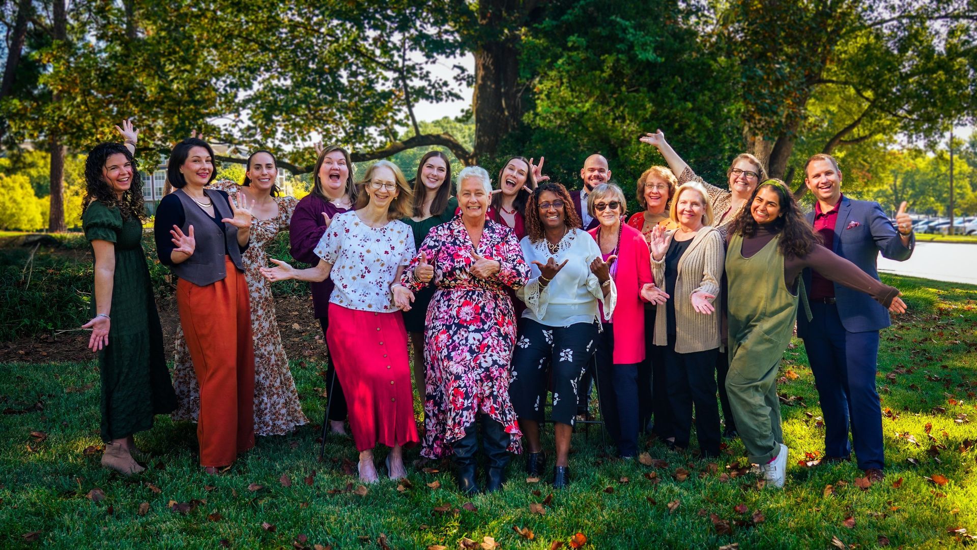 Group of people standing outdoors in a garden, smiling and raising their arms in colorful outfits.