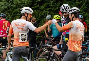 Cyclists in helmets high-five at a race start, with bikes and orange-white jerseys in the foreground.
