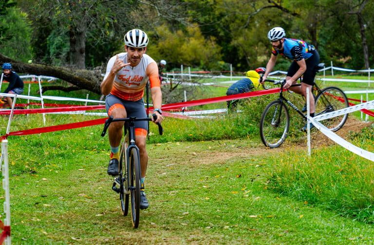 Two cyclists race on a grassy course, one in orange leading and another in blue navigating a red-and-white barrier.