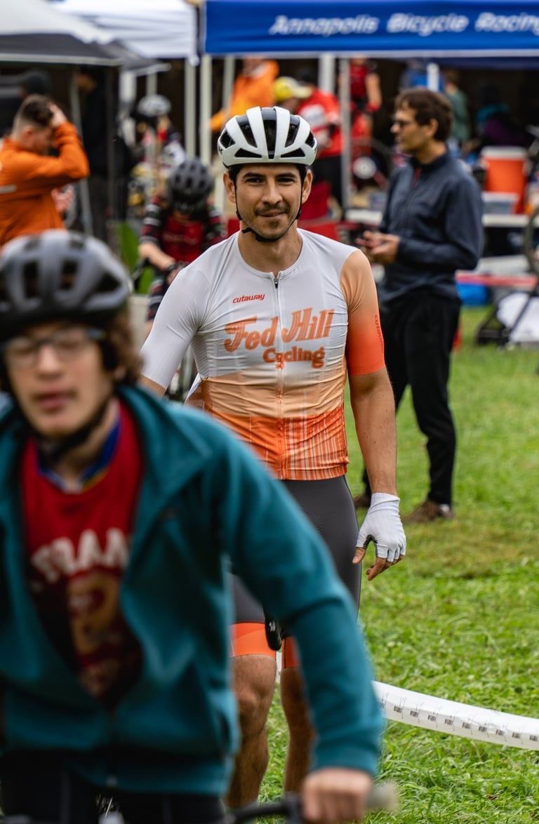 Cyclist in white-and-orange jersey standing on a grassy race course with spectators behind him