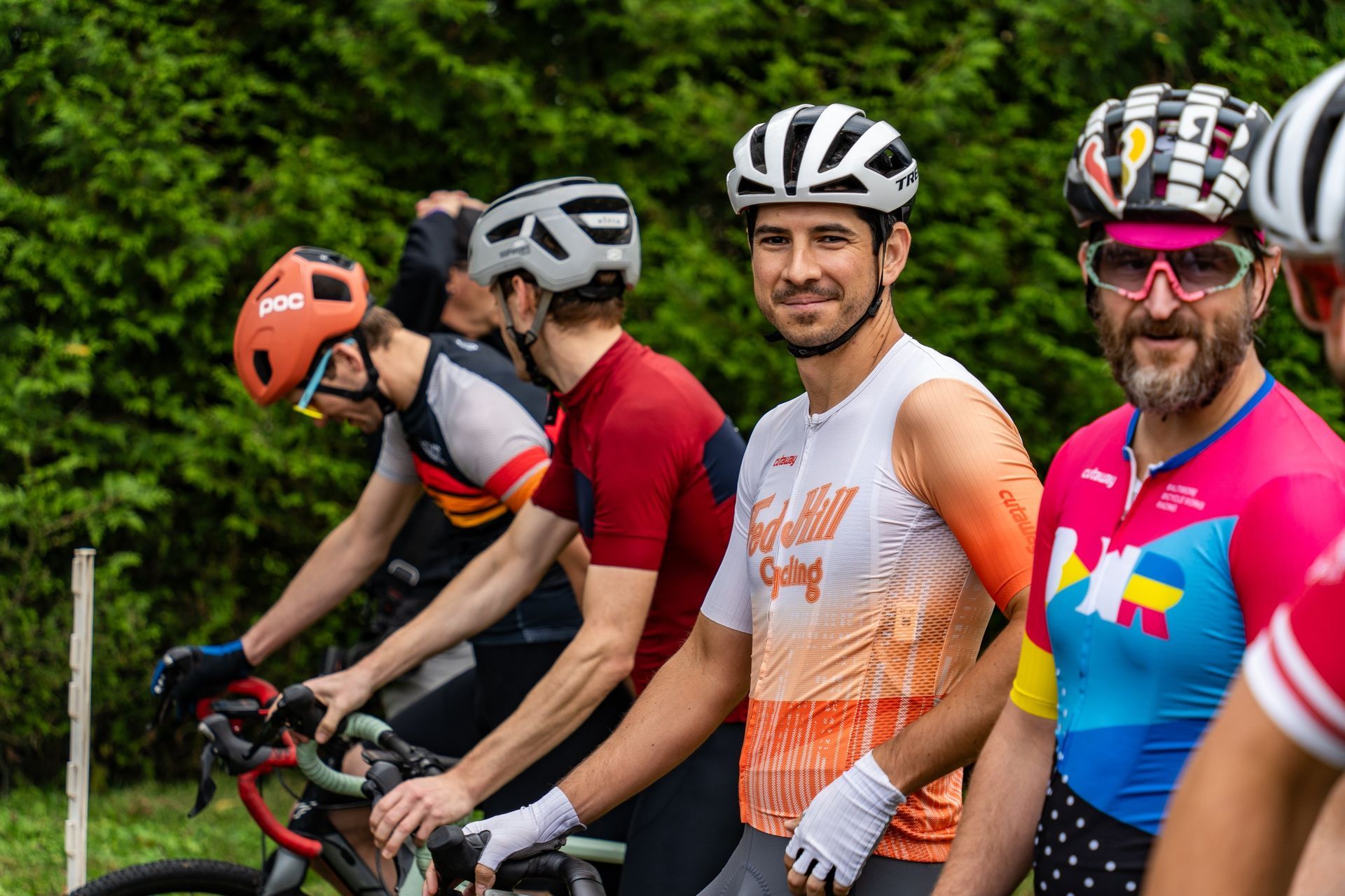 Cyclists lined up on bikes outdoors, wearing colorful helmets and jerseys, smiling at the camera