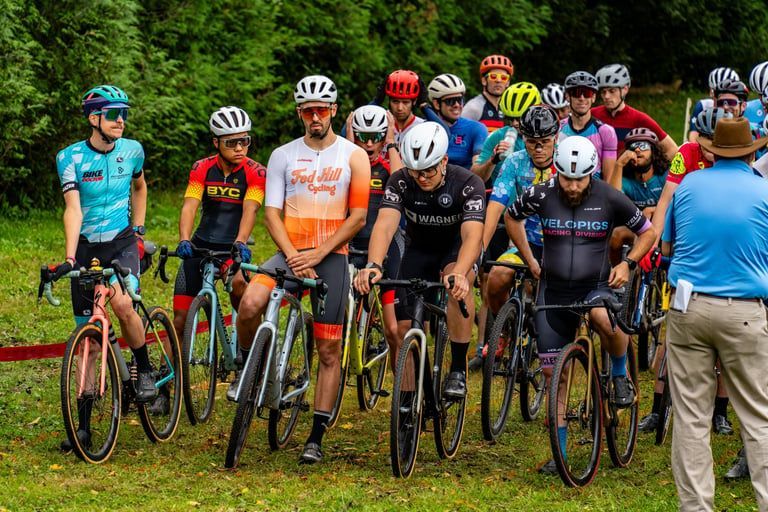 Cyclists lined up at a grassy start line, wearing colorful jerseys and helmets before a race.