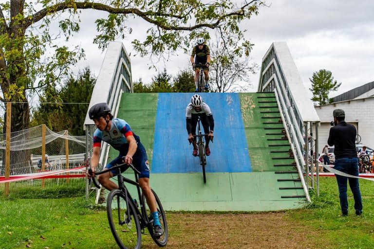 Cyclists riding a blue-green ramp obstacle course in a grassy park, with one rider jumping over the center ramp
