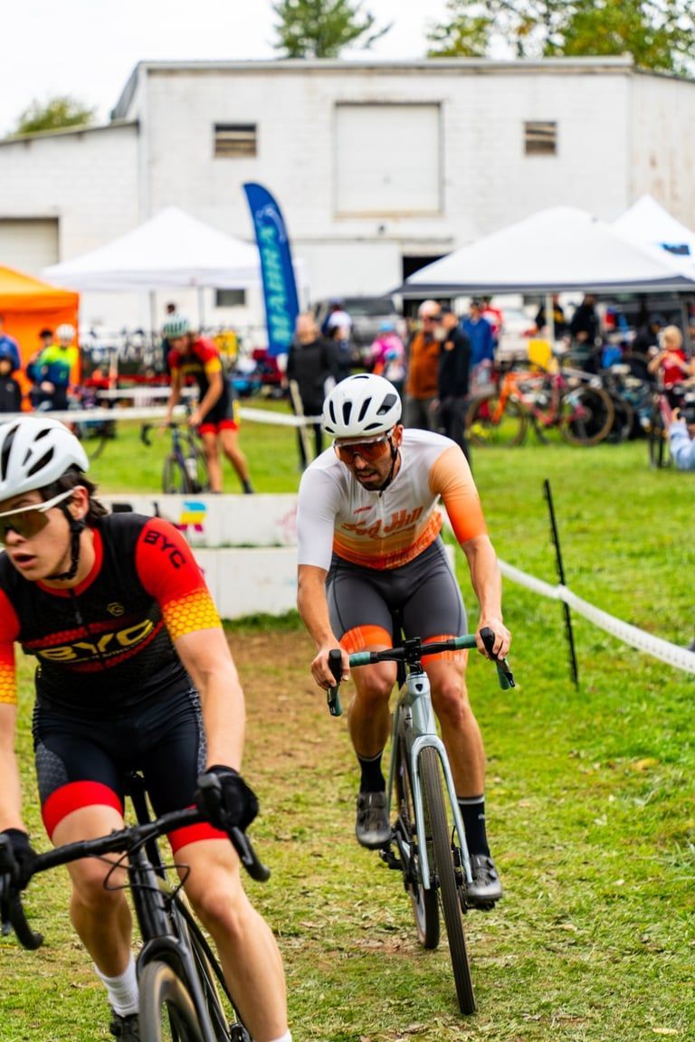 Cyclists racing on a grassy course near tents and spectators, with one rider in the foreground.