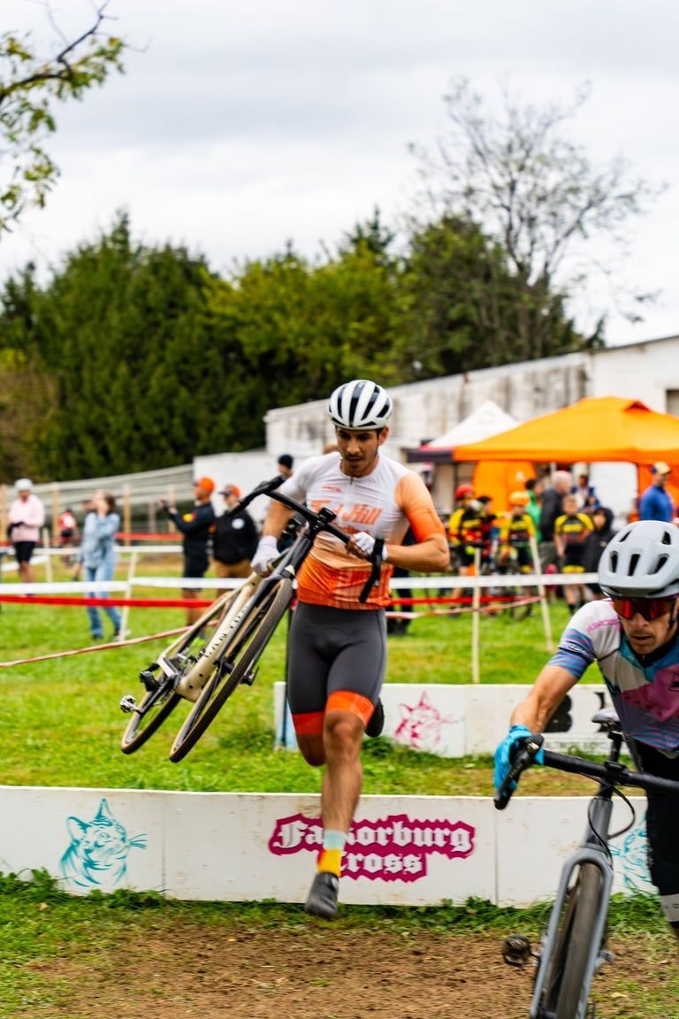 Cyclocross rider carrying a bike over barriers during a race, with orange tents in the background.