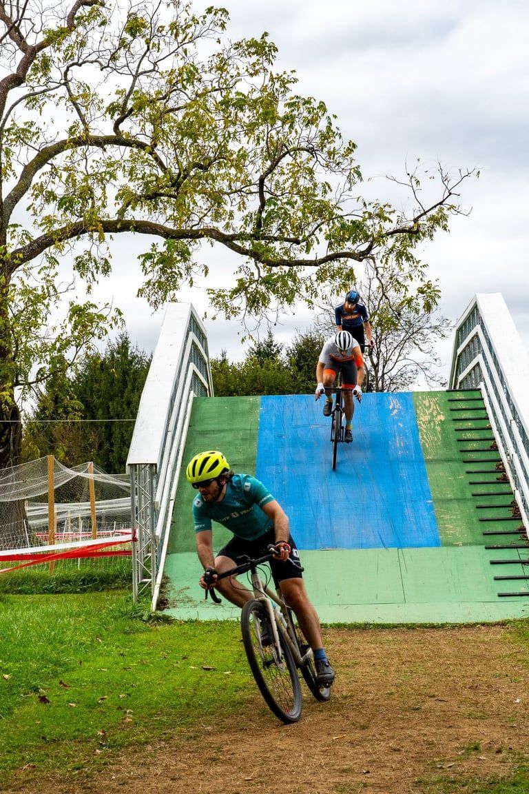 Cyclists riding down a blue-green ramp on a grassy outdoor course, one turning at the bottom