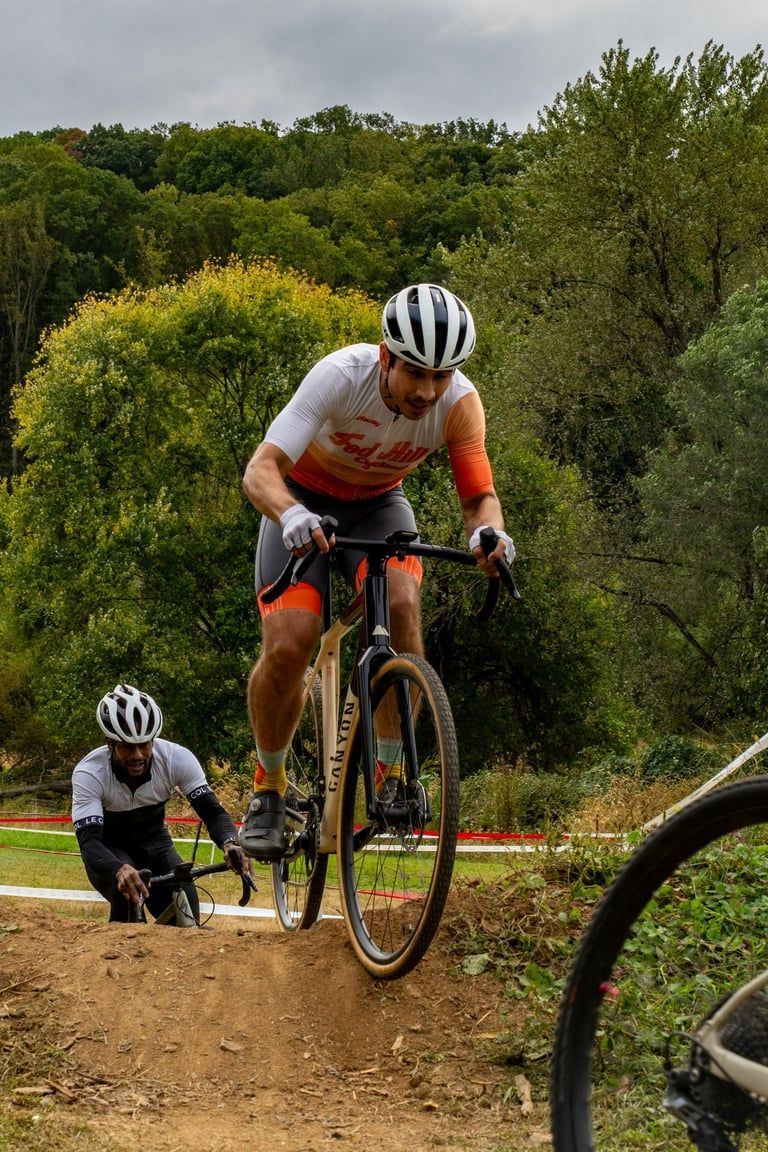 Two mountain bikers race on a dirt trail through a wooded area, one airborne over a small jump.