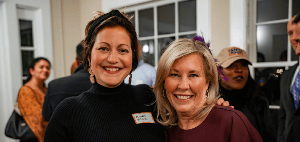 Two smiling women stand together indoors at an event, with people visible in the background.