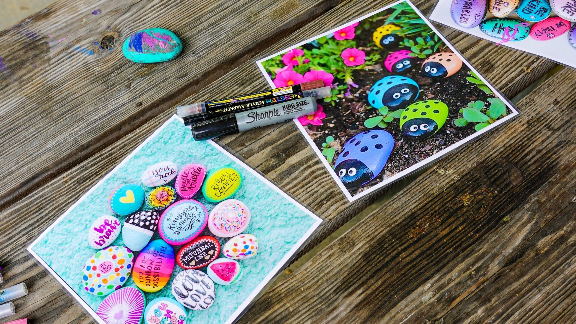 Colorful painted rocks with smiling faces and hearts on a wooden table, with markers and a decorated card nearby