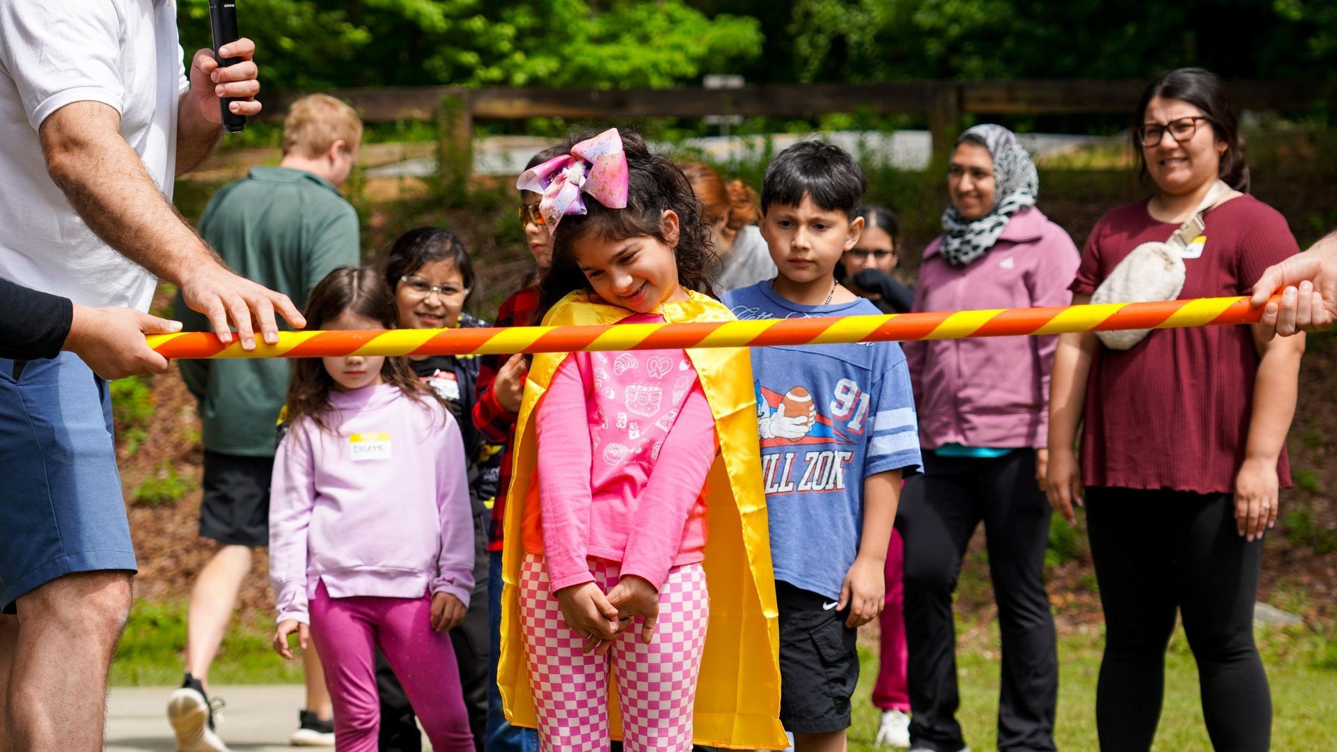 Children and adults at an outdoor ribbon-cutting ceremony in a park, holding a bright orange ribbon