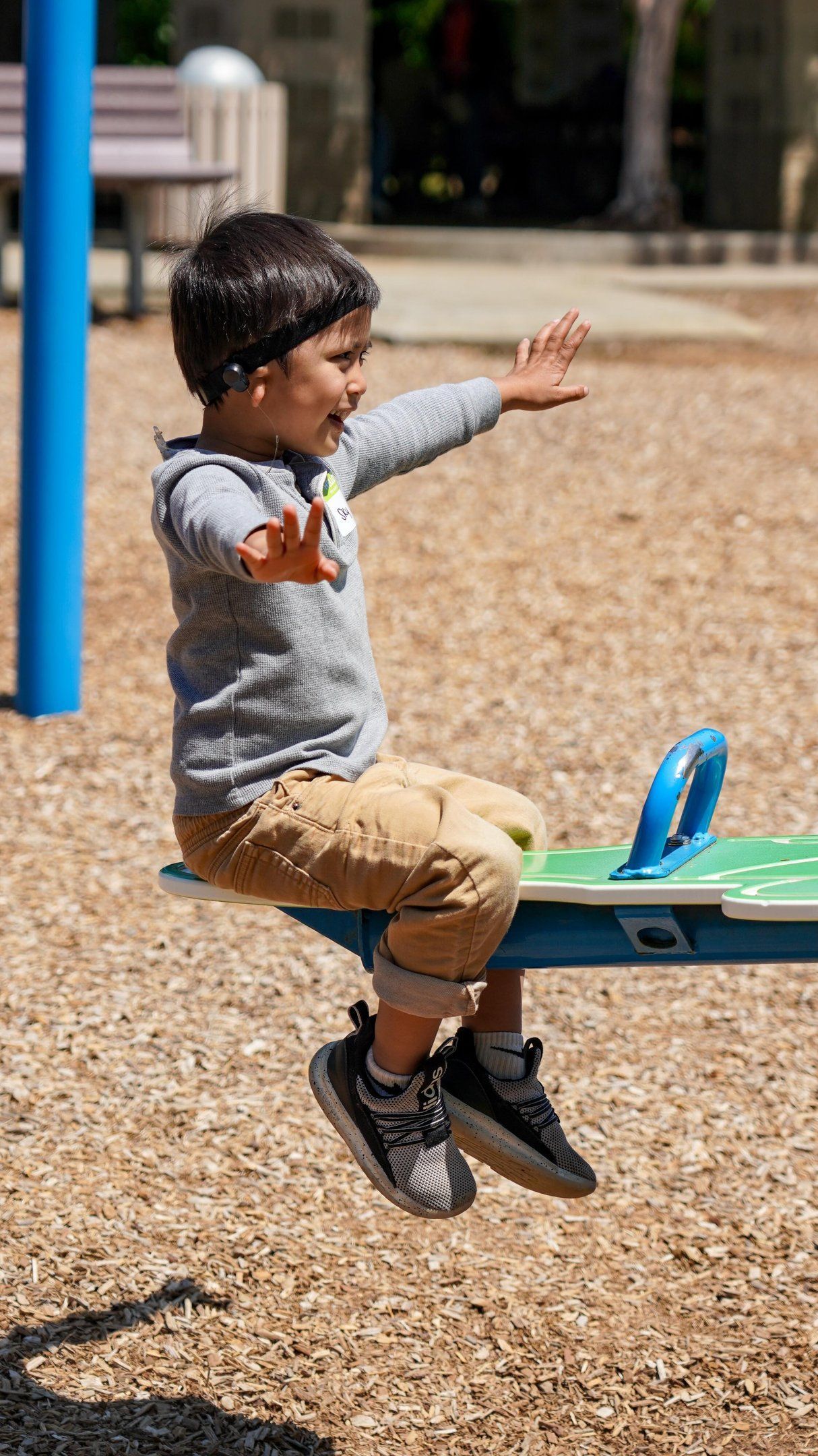 Child balancing on a playground seesaw, arms outstretched, wearing a gray sweater and tan pants.