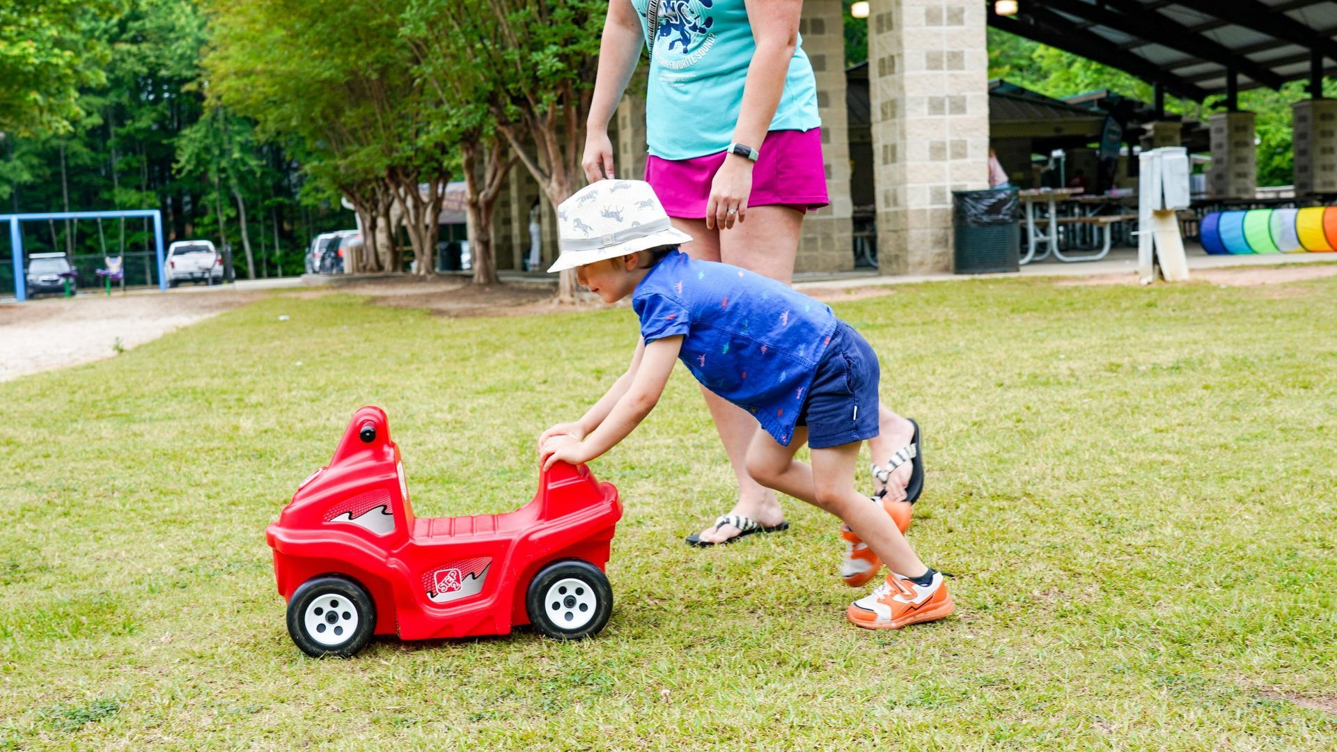 Child in blue shirt pushes a red toy car on grass near an adult outdoors