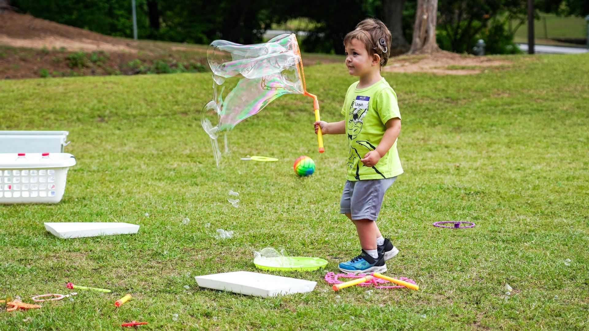 Child blowing a large soap bubble in a grassy park with bubble trays and toys nearby