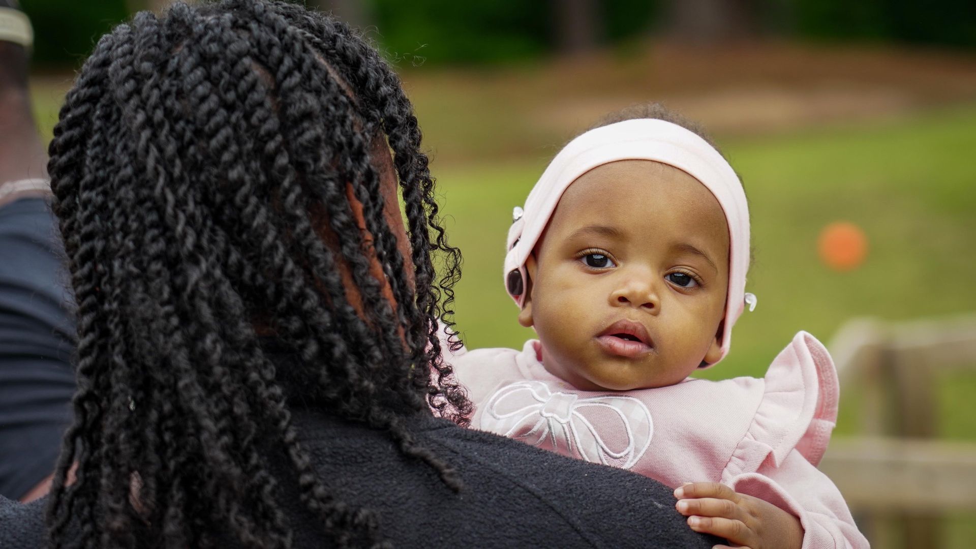 Woman holding a baby outdoors; the baby wears a pink outfit and white headband.