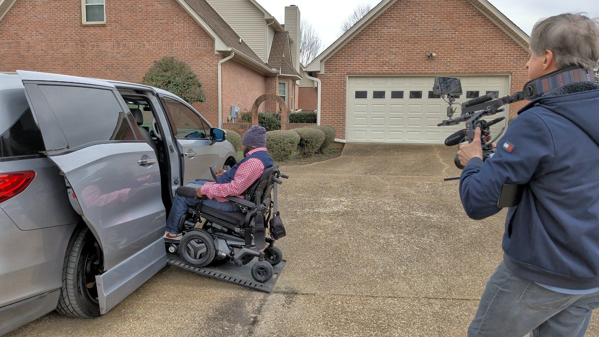 Person in a wheelchair exiting a silver van while a cameraman films in a suburban driveway