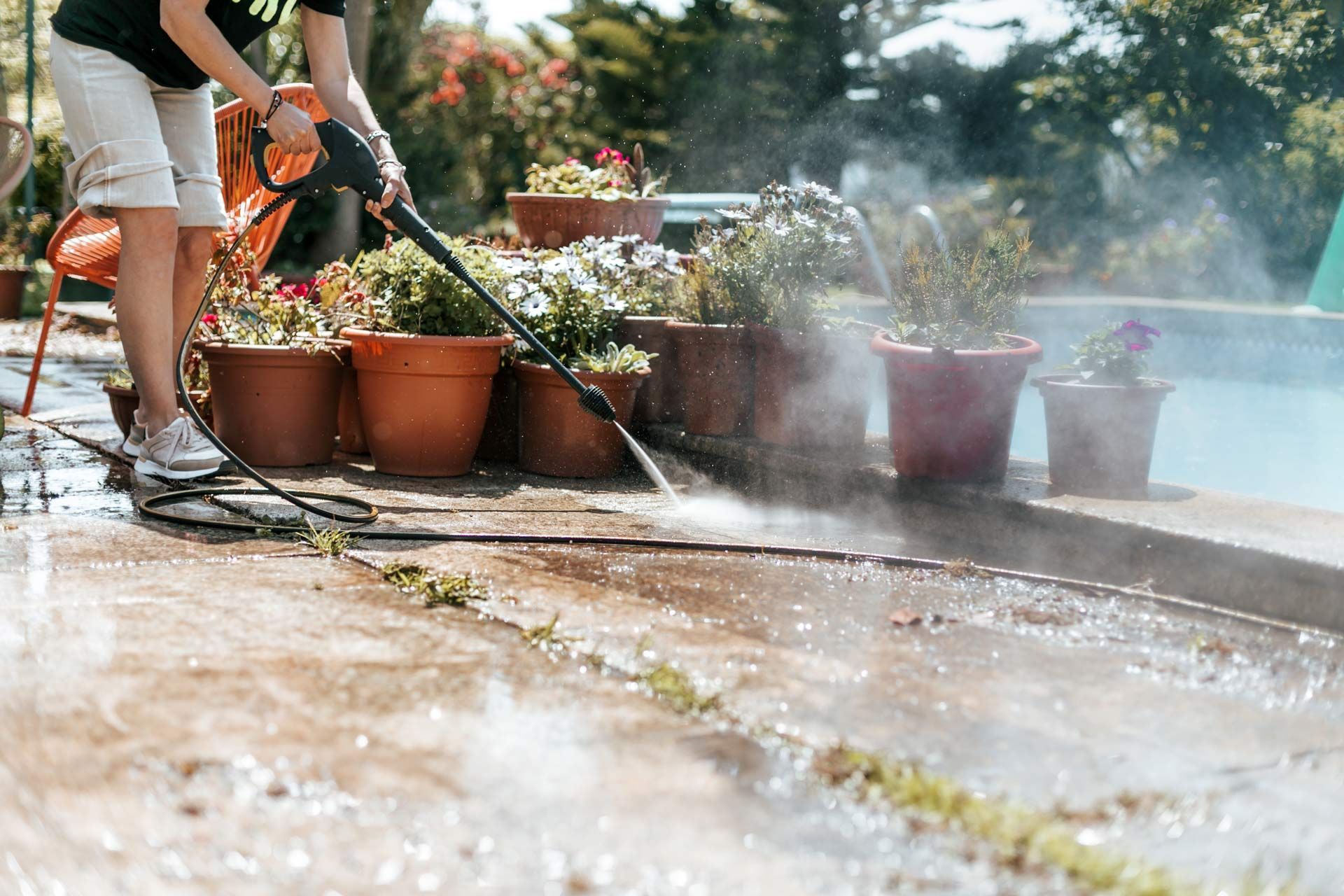 Person pressure washing a concrete patio near potted plants and a pool.