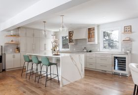 A modern kitchen featuring white cabinets, a marble-topped island with three green stools, and light wood flooring.