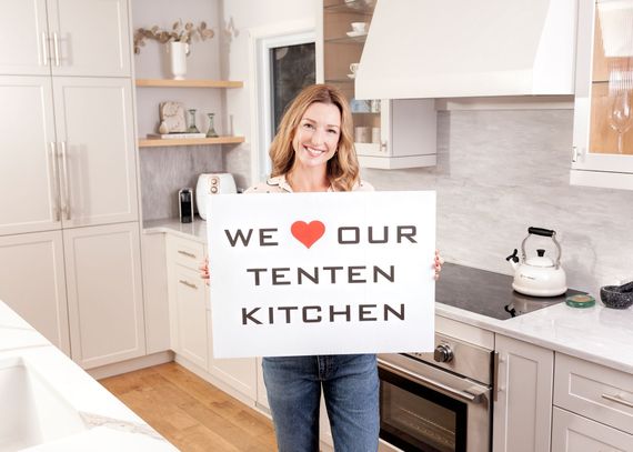 A smiling person in a bright kitchen holds a sign that reads