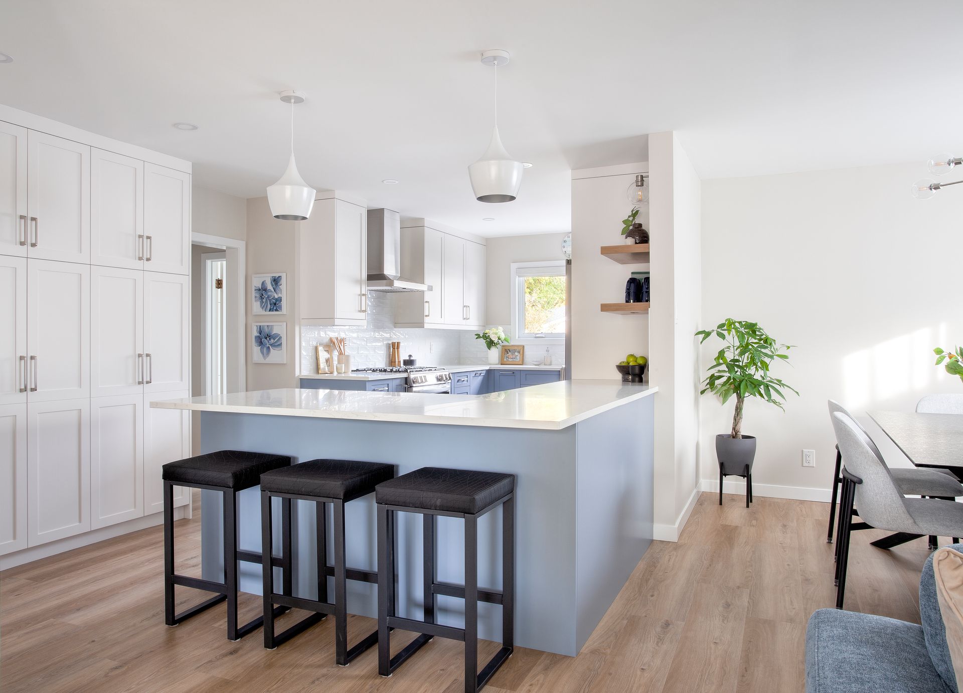 Modern kitchen with a light blue island, three dark barstools, white cabinetry, and a dining area with a potted plant.