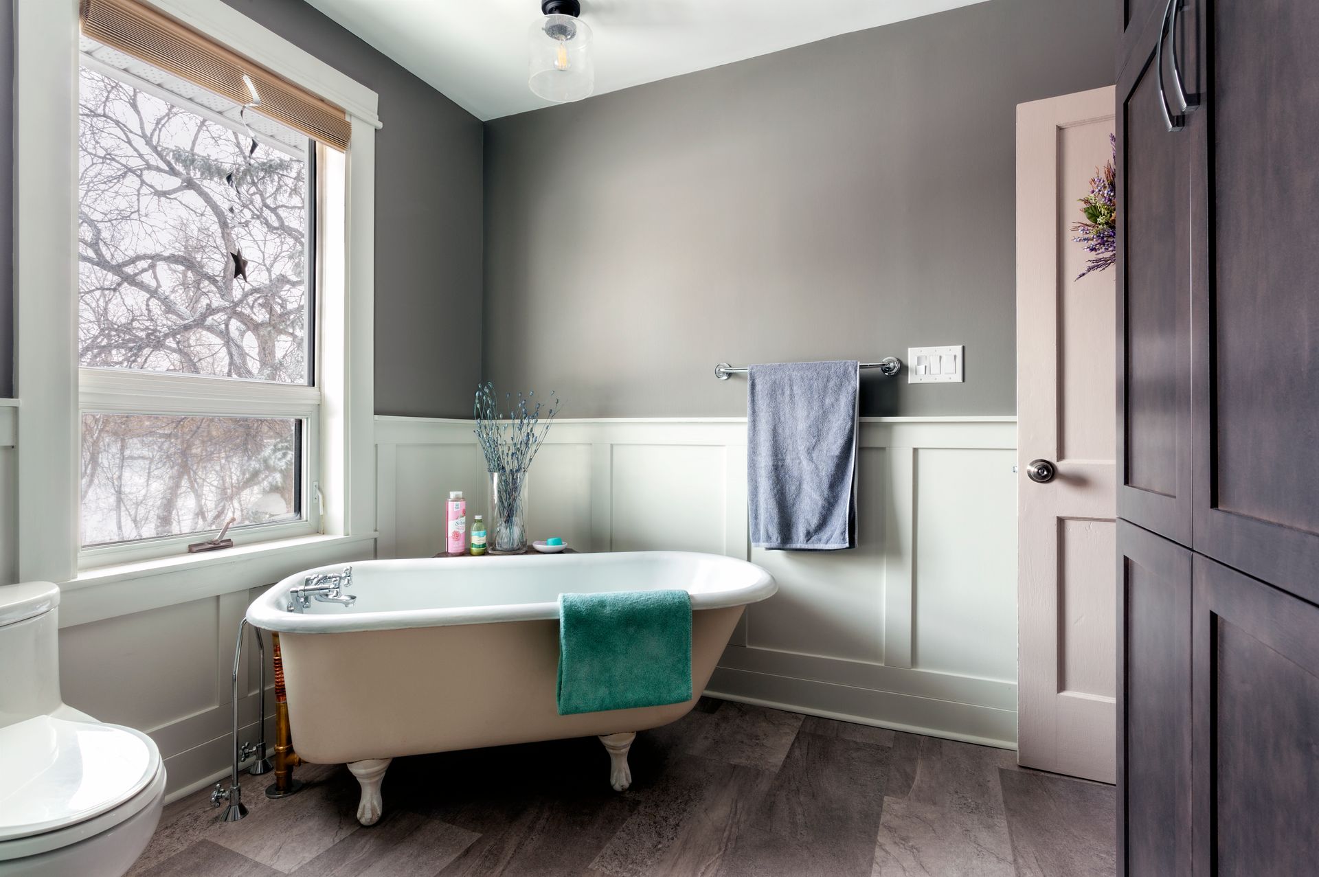 A clawfoot tub sits in a bathroom with white wainscoting, gray walls, a window with a tree view, and dark wood cabinetry.