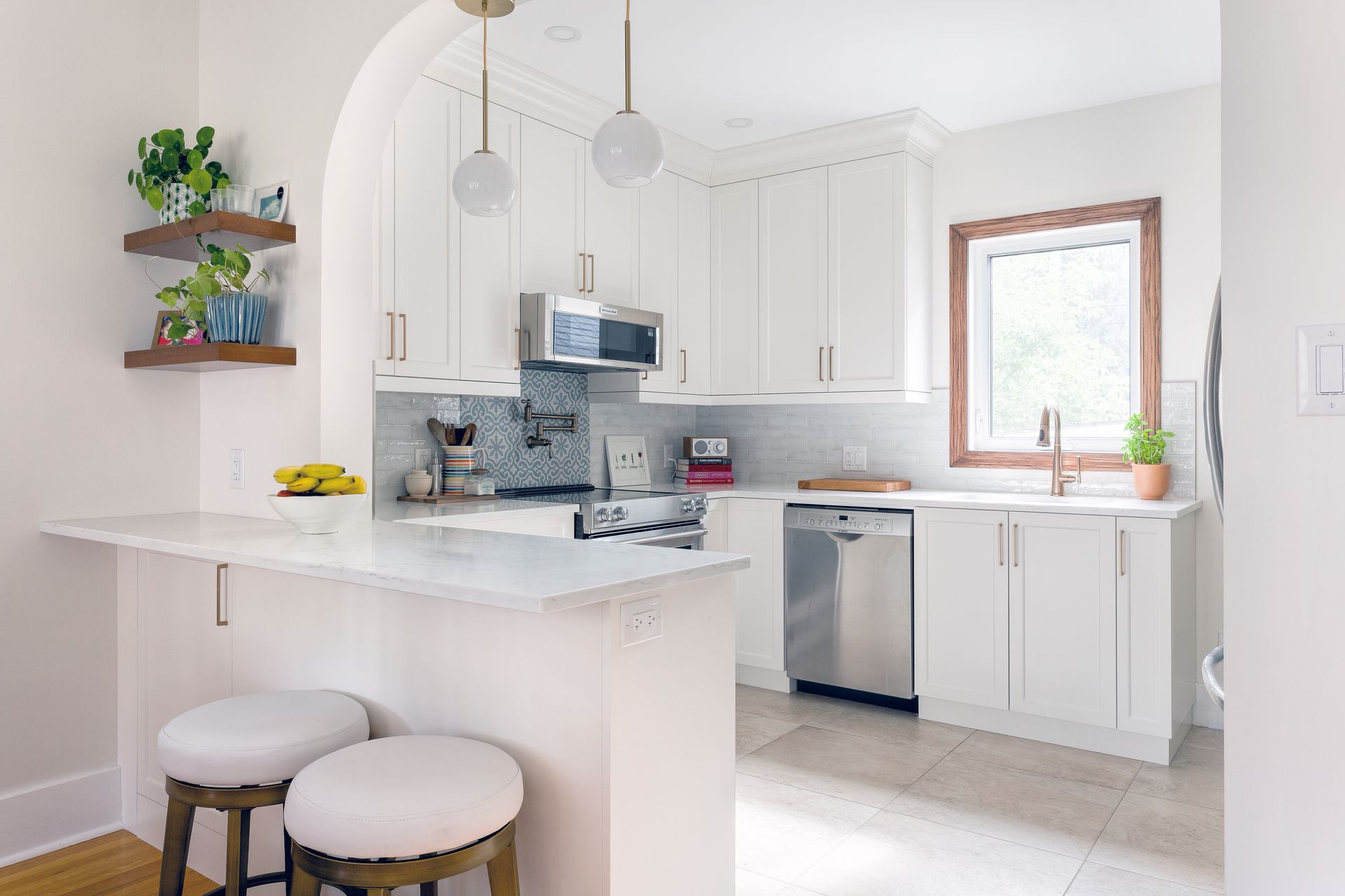 A bright, modern kitchen featuring white cabinets, stainless steel appliances, a white marble peninsula, and two bar stools.