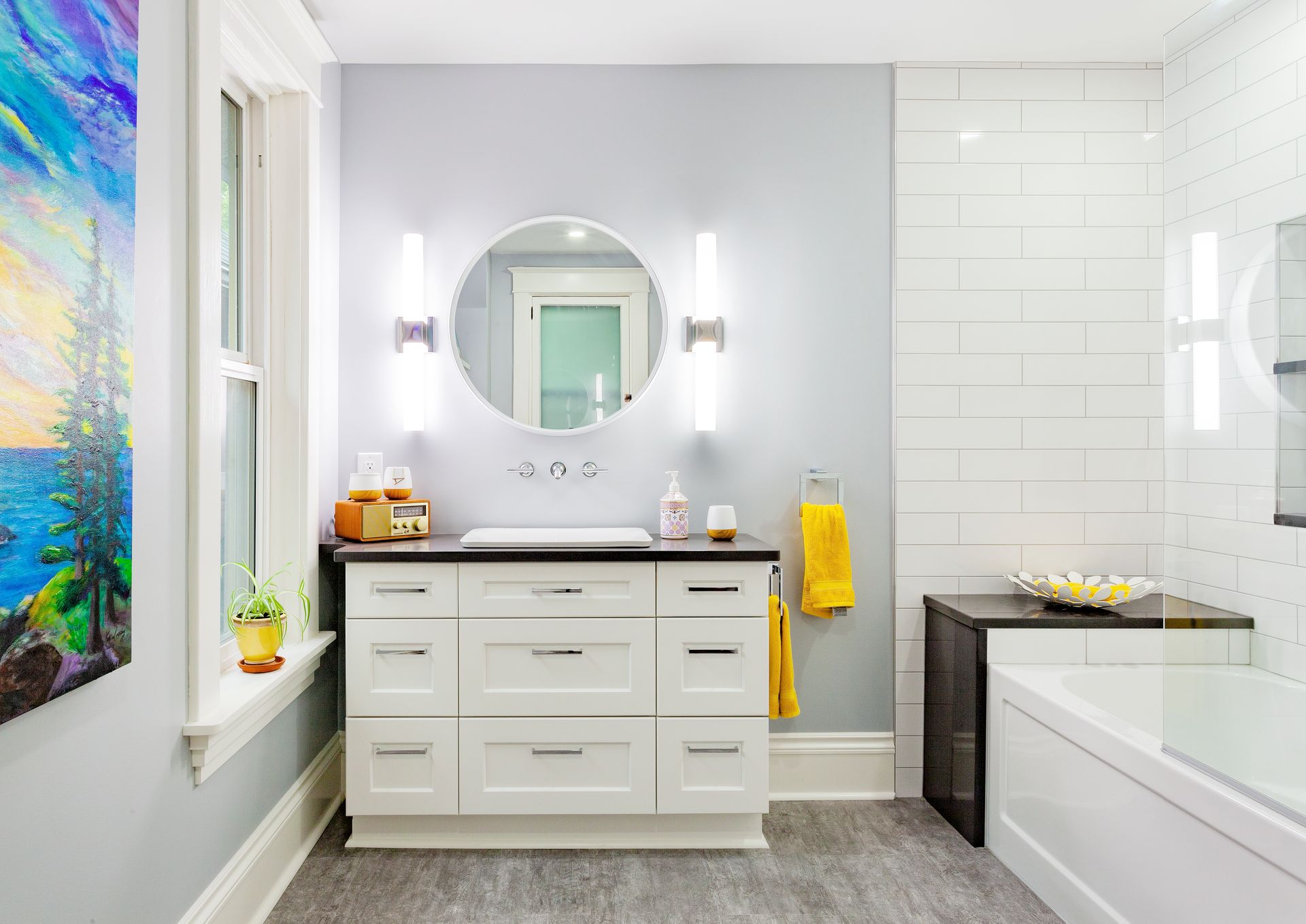 Modern bathroom with a white vanity, round mirror, light gray walls, white subway tile shower, and a vibrant wall painting.