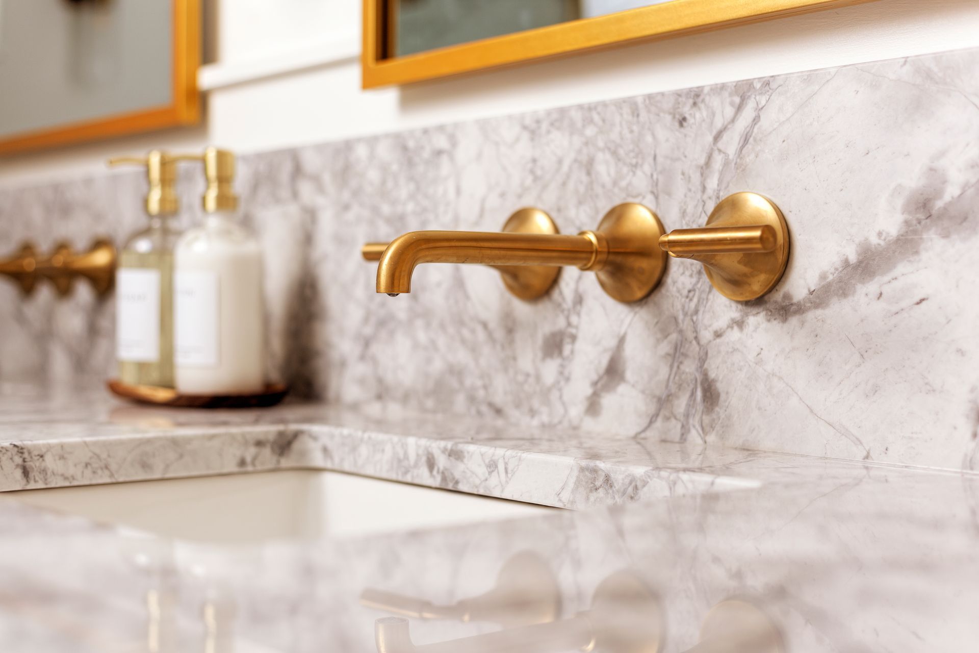 Wall-mounted brass faucet over a white marble countertop with a matching sink and soap dispensers.