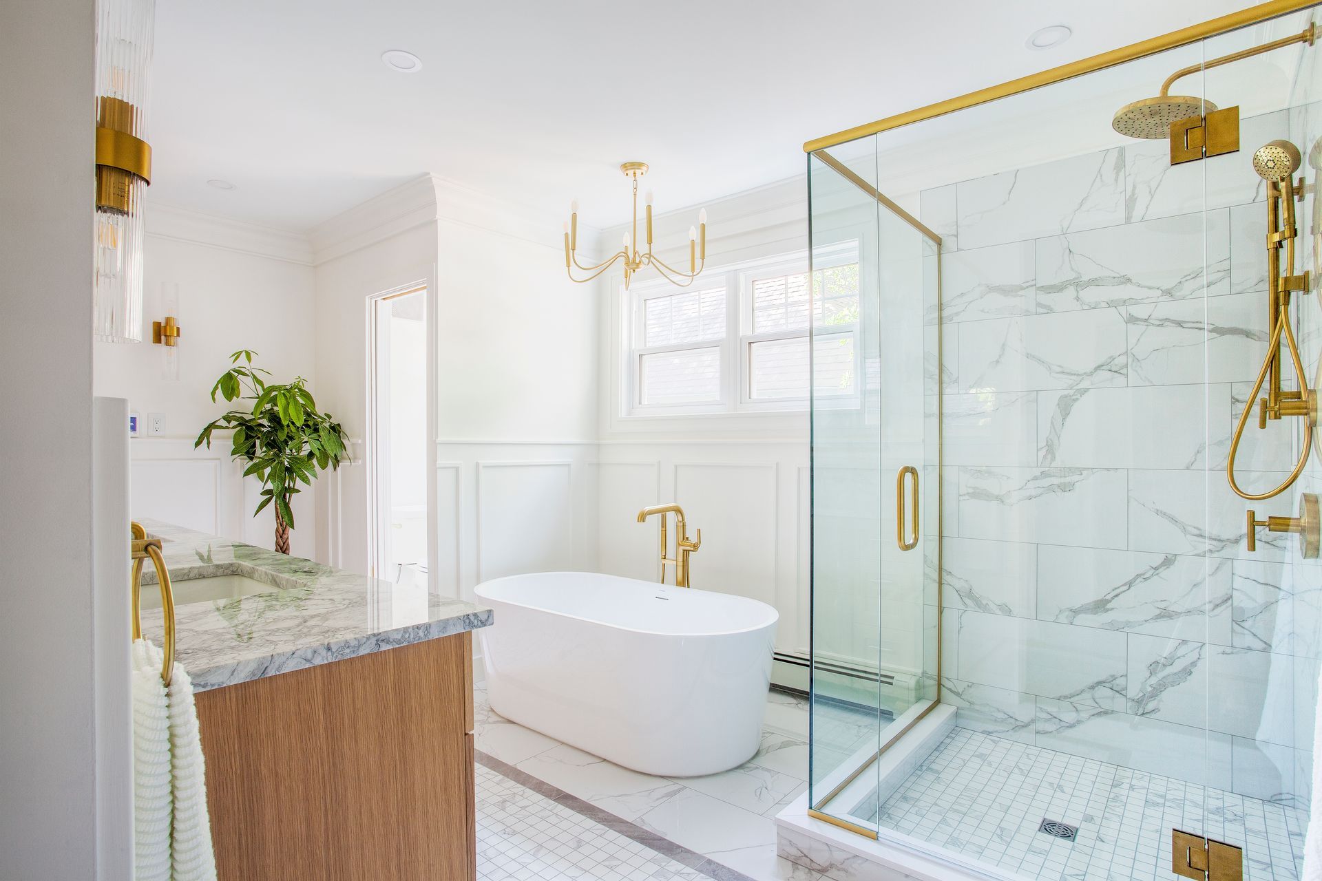 A bright, modern bathroom with a white freestanding tub, glass walk-in shower with gold fixtures, and marble flooring.