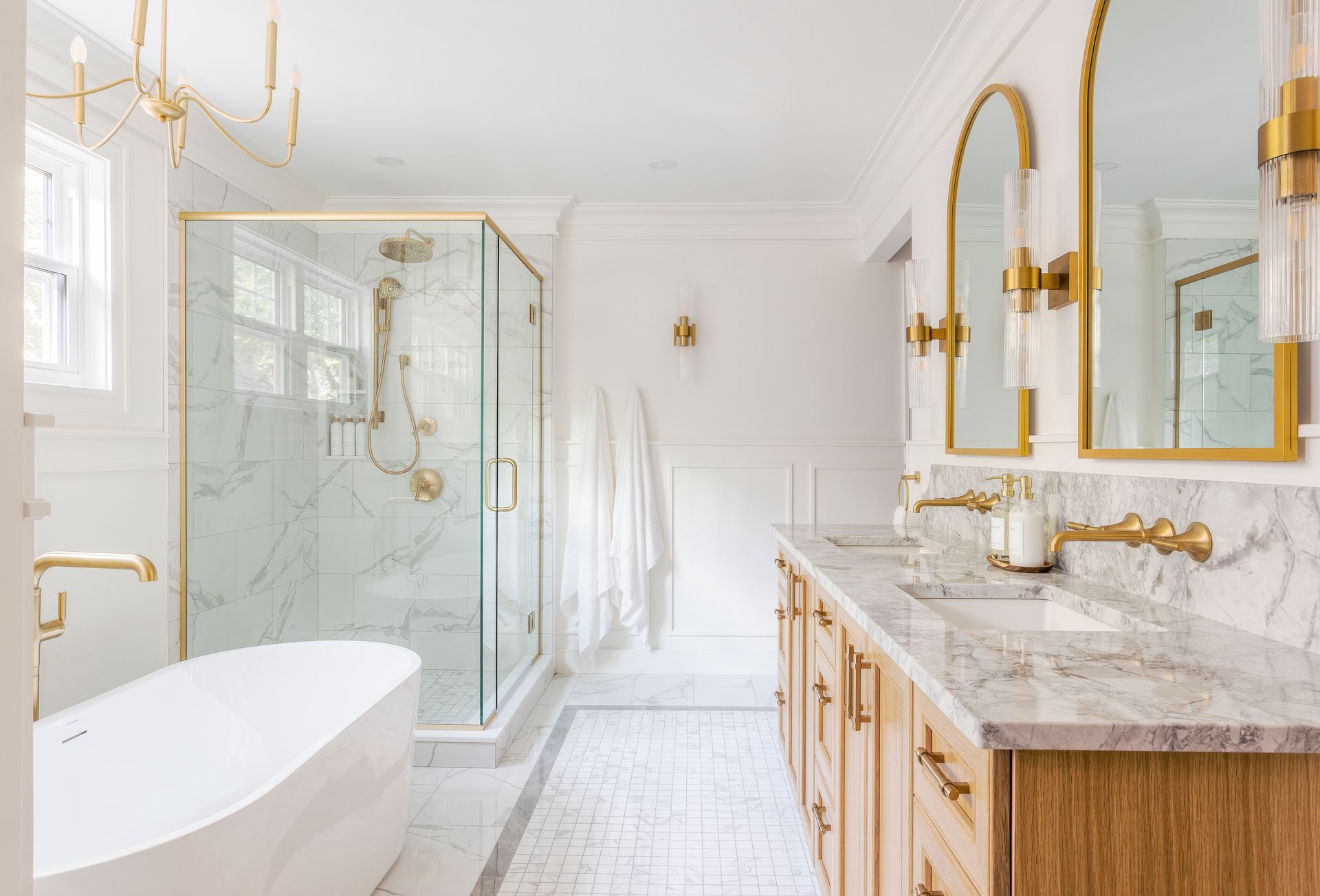 A modern bathroom with a white freestanding tub, a glass walk-in shower, a wooden double vanity, and gold fixtures.