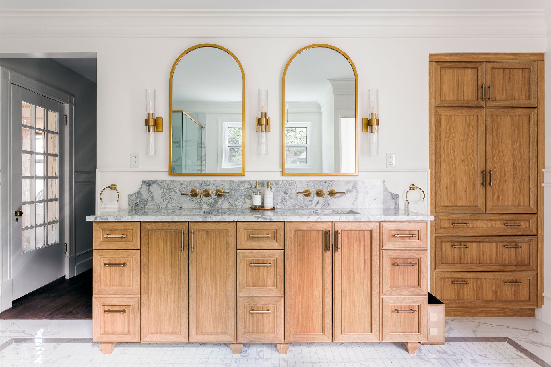 A double vanity bathroom with light wood cabinets, gold fixtures, arched mirrors, and marble countertops.