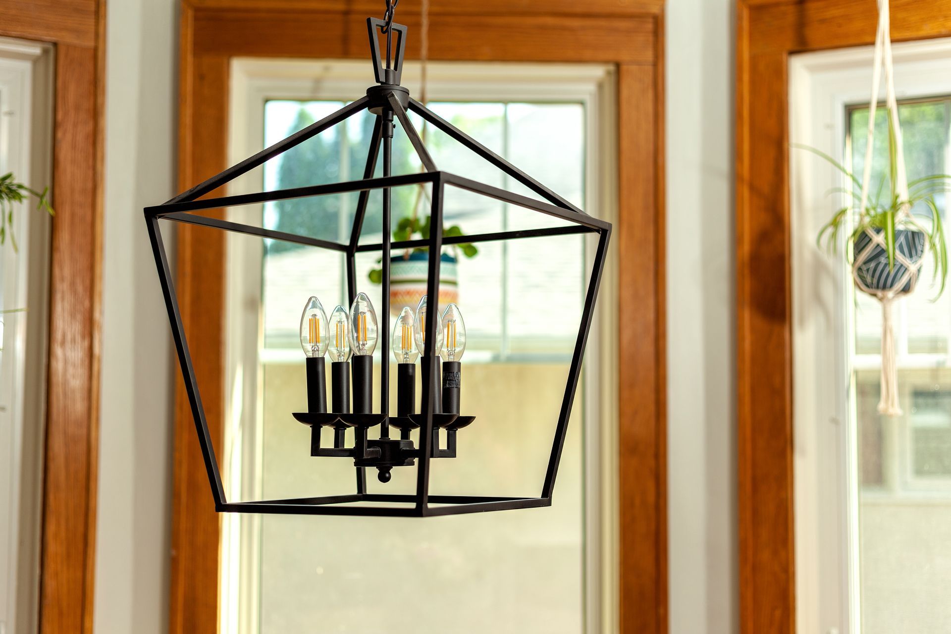 A black geometric, four-light metal pendant chandelier hangs in front of a window with wooden trim.