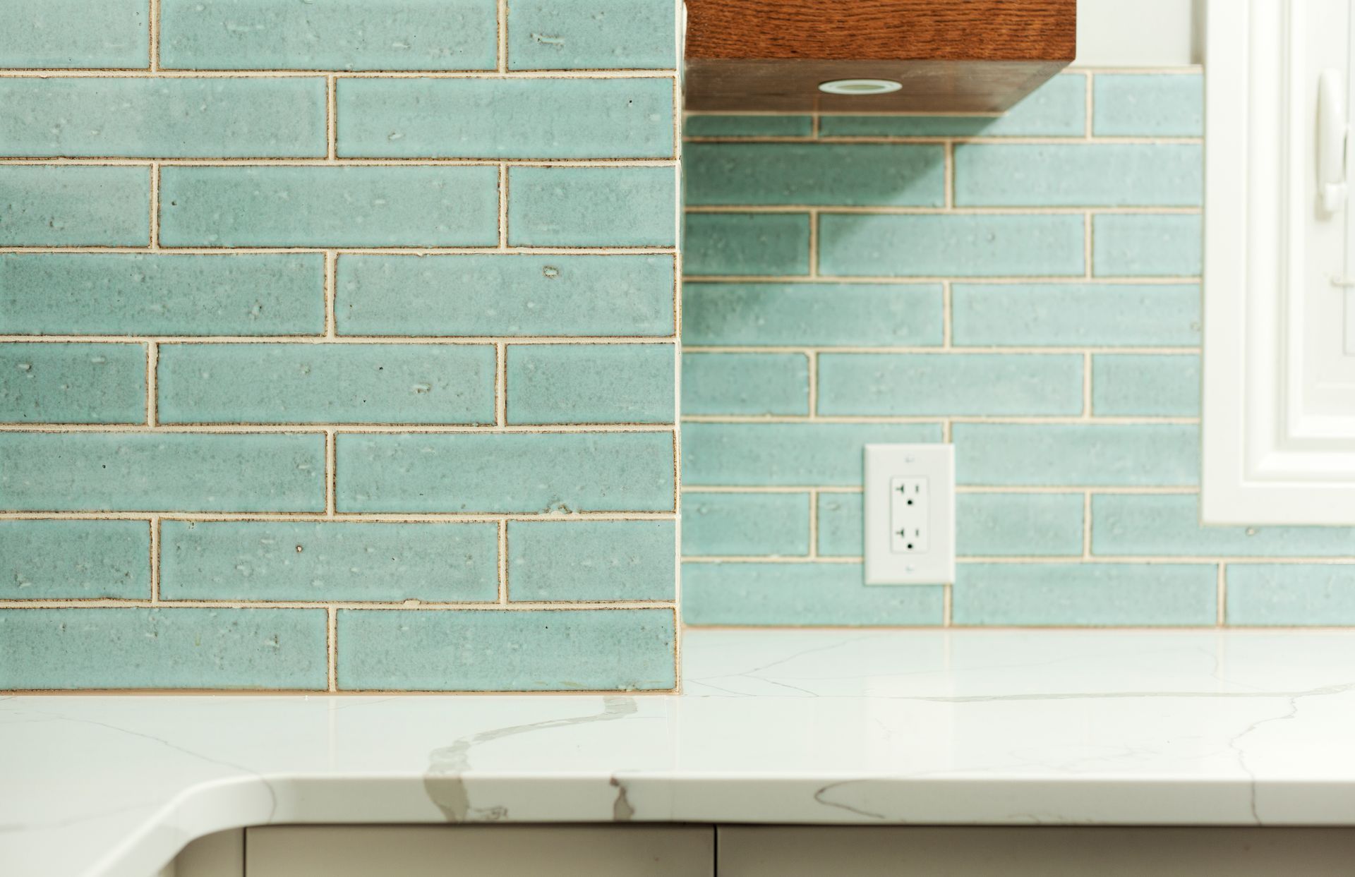 A kitchen backsplash featuring light blue rectangular tiles and white marble countertops with a power outlet.