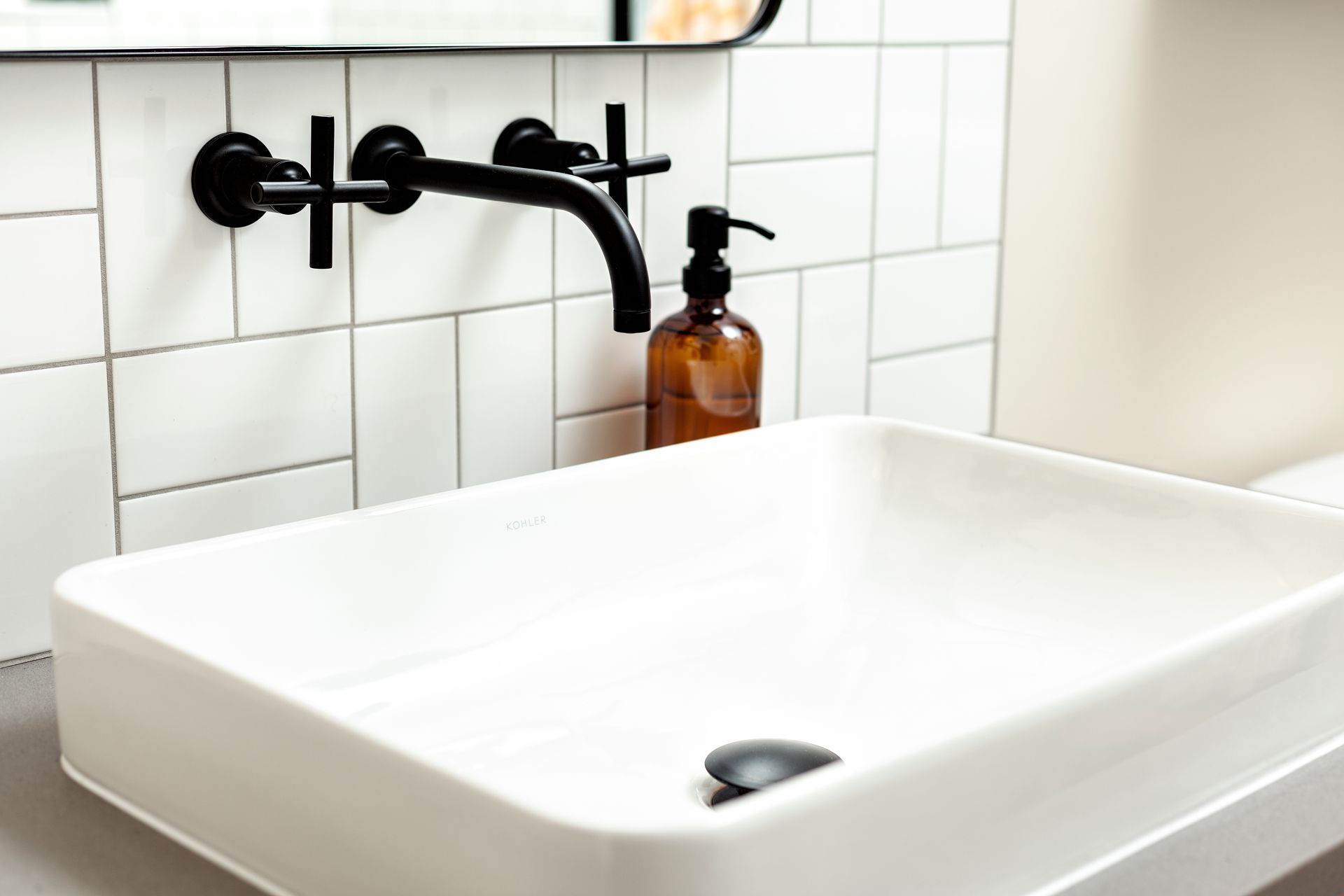 A white rectangular vessel sink with a black wall-mounted faucet and a brown soap dispenser against white subway tiles.