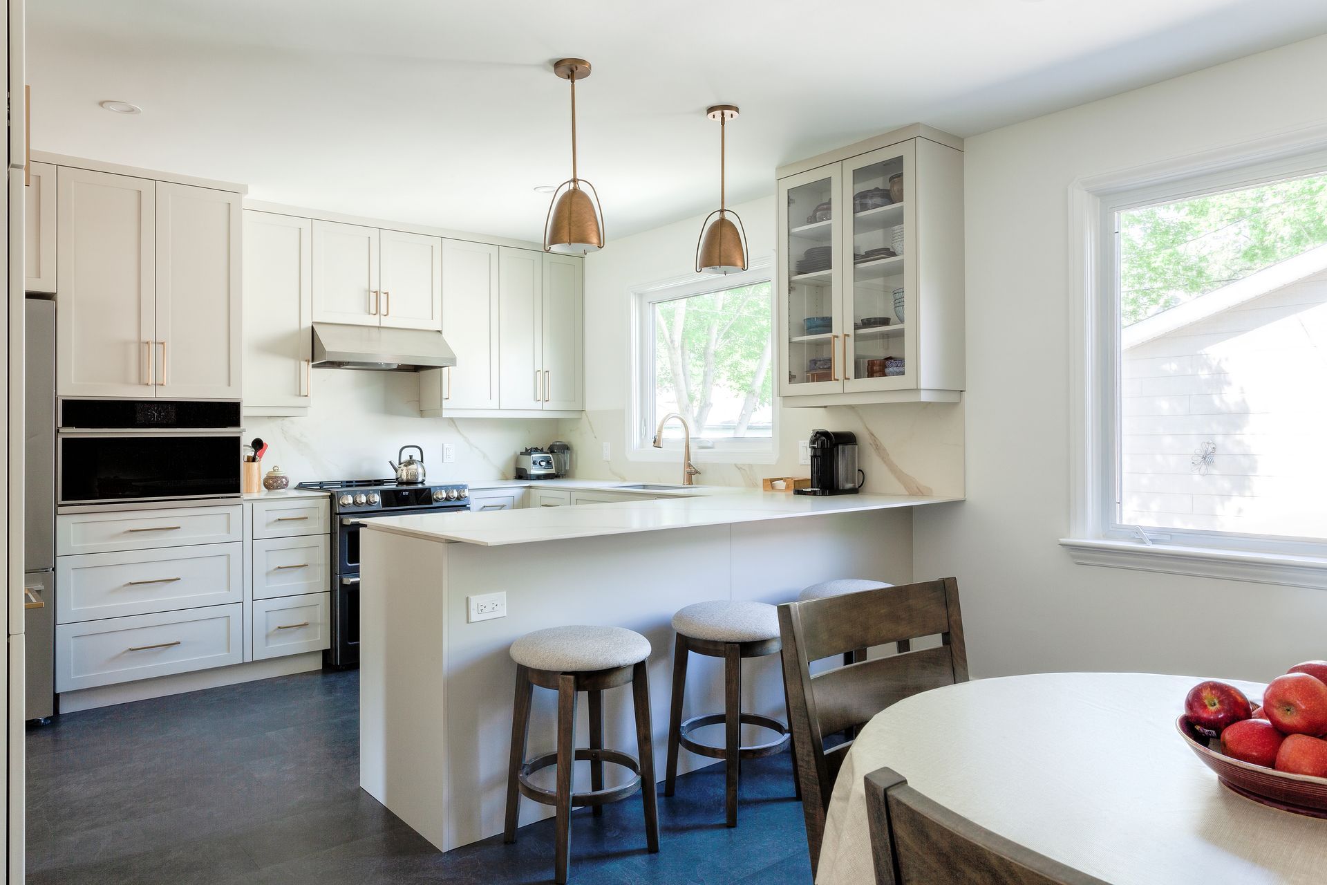 A bright, modern kitchen featuring white cabinets, a stone island with two stools, and a small dining table with apples.