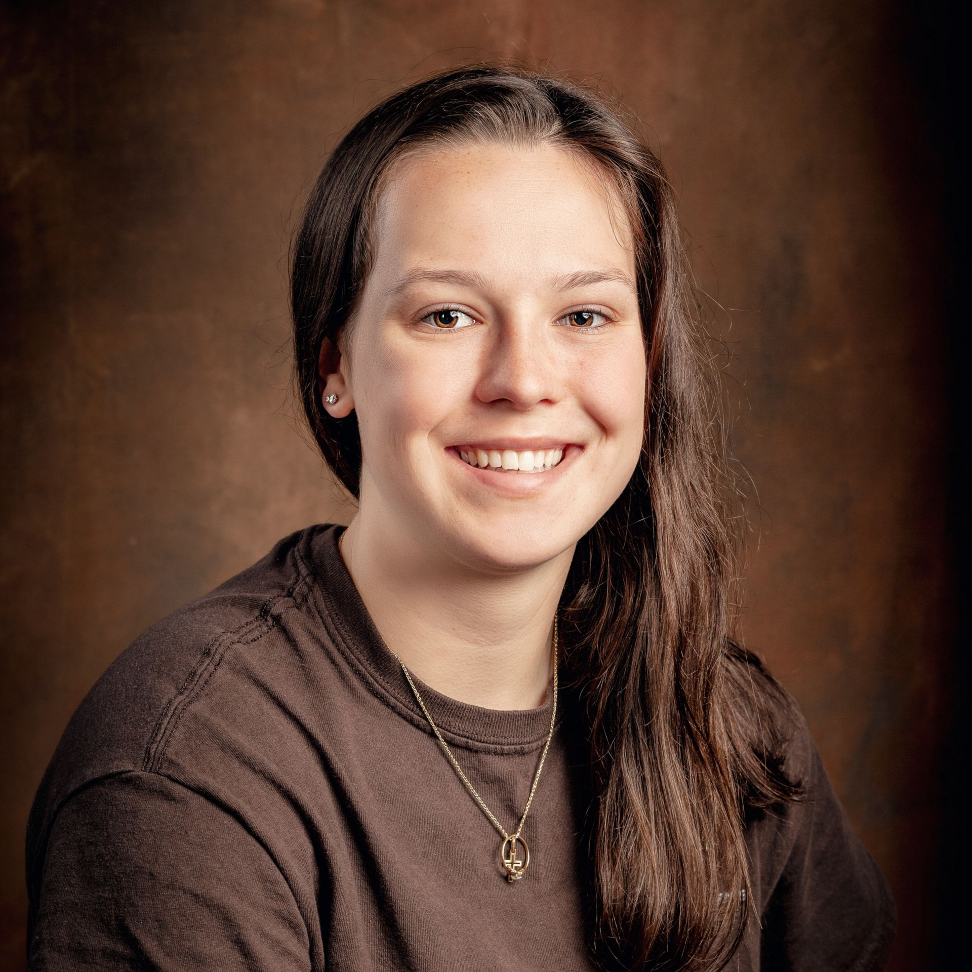 A smiling person with long brown hair, wearing a brown t-shirt and a necklace, posed against a dark brown background.