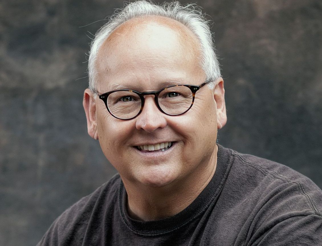 A smiling person with gray hair and glasses wearing a dark brown sweater against a neutral, blurred background.