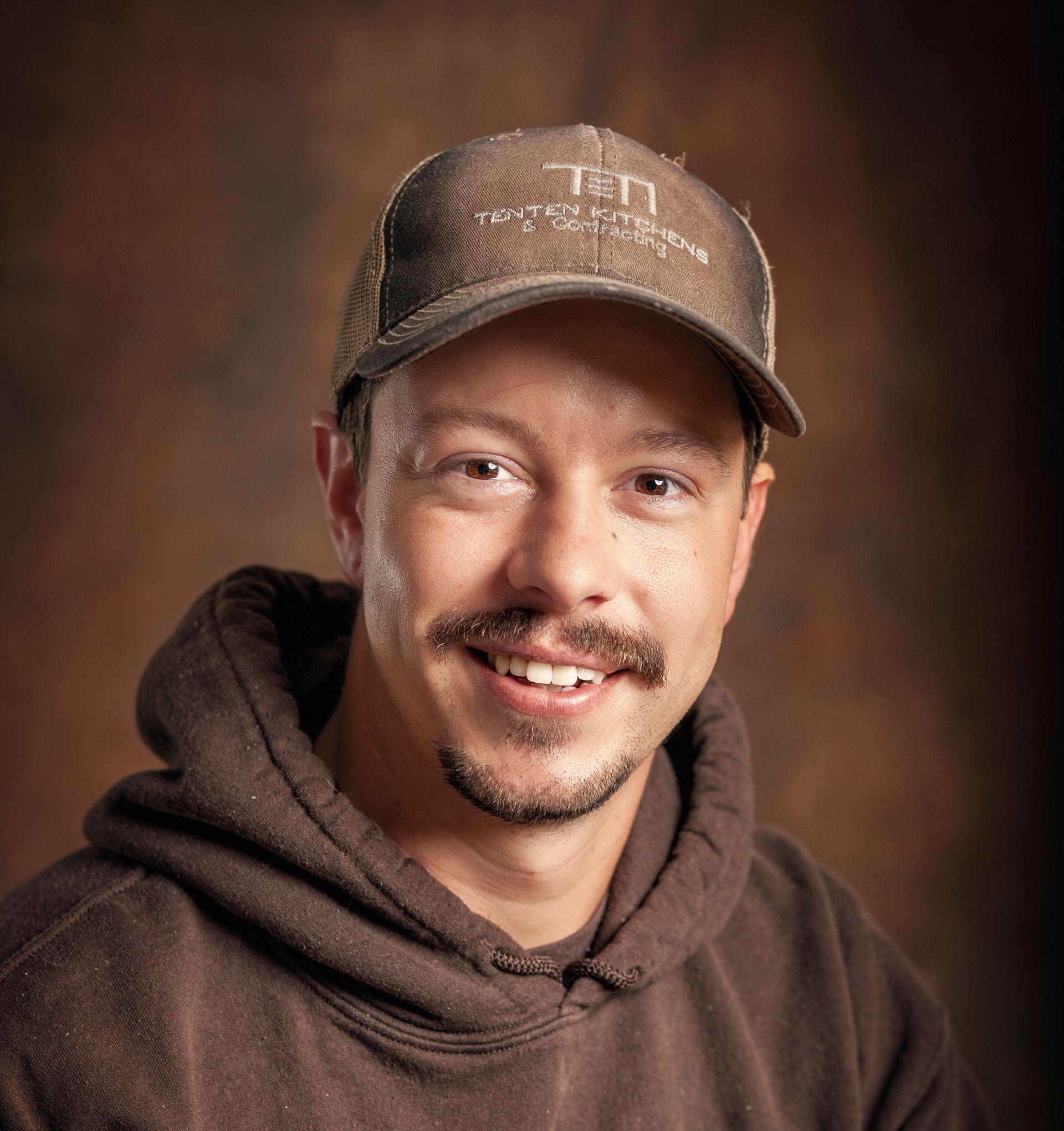 A smiling person wearing a brown cap and brown hoodie against a dark, mottled background.