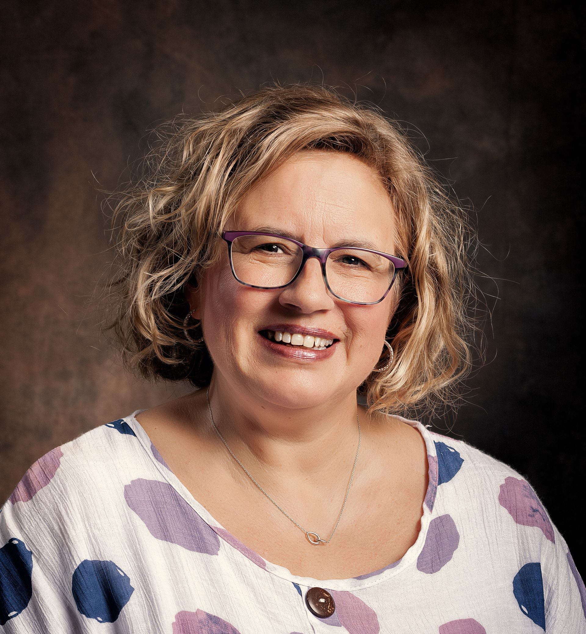 A smiling person with curly hair and glasses, wearing a white shirt with blue and purple polka dots against a dark backdrop.