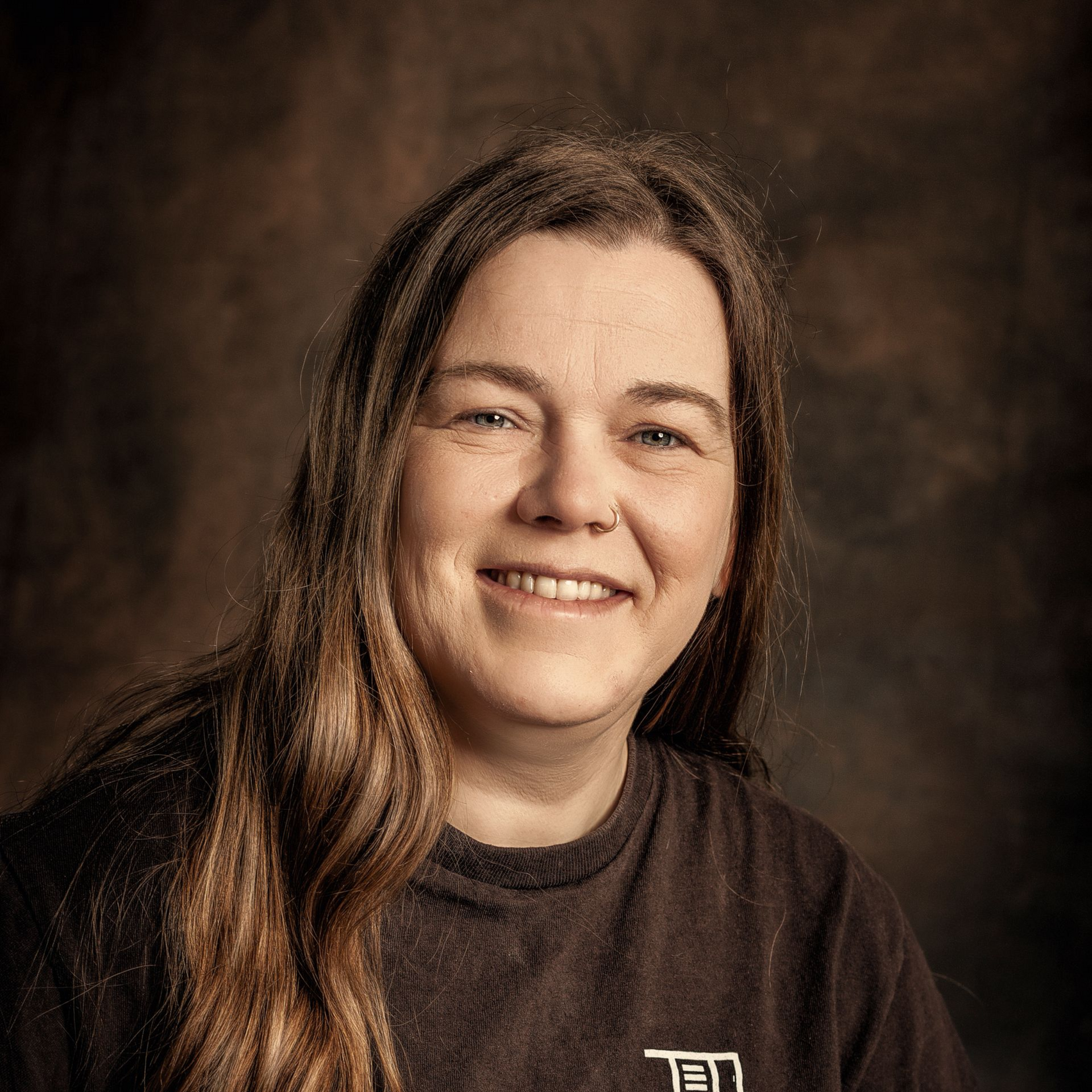Smiling person with long brown hair in a dark T-shirt against a brown studio backdrop