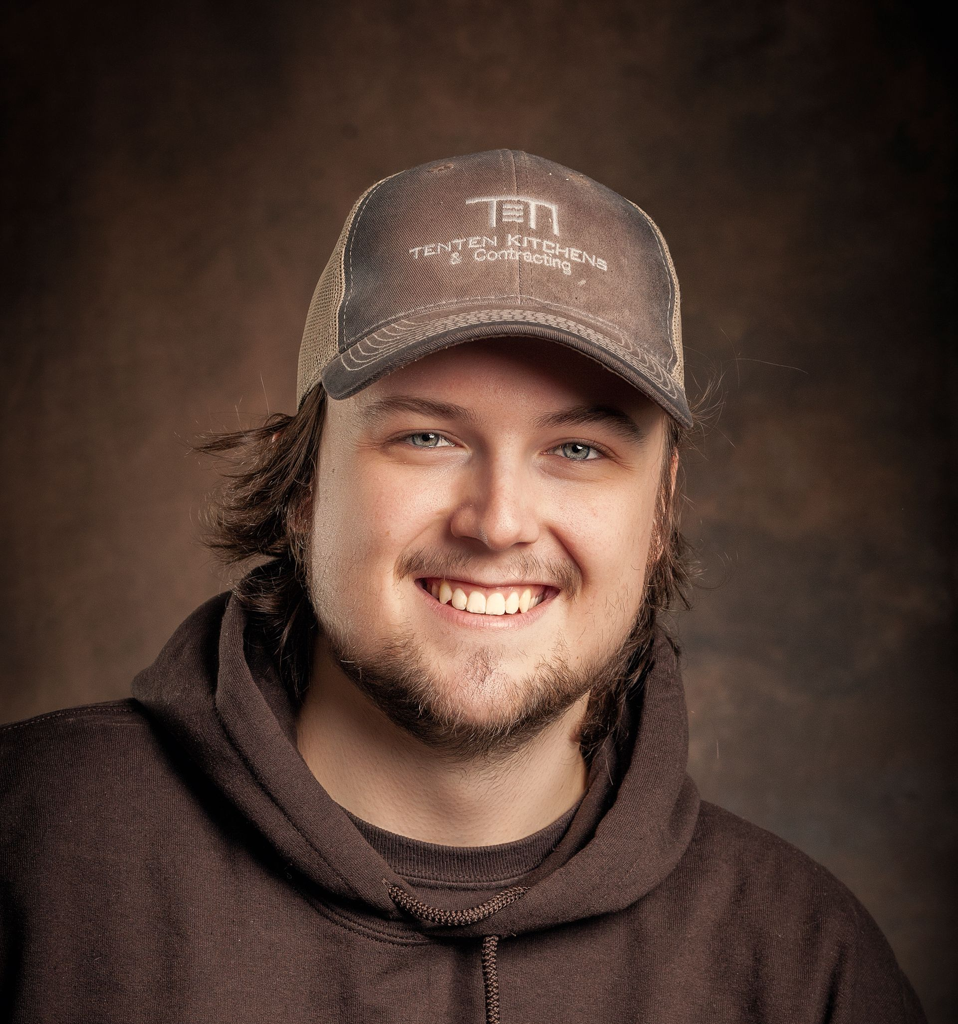 Smiling person with brown hair, wearing a brown hoodie and a logo-branded baseball cap against a dark, textured backdrop.