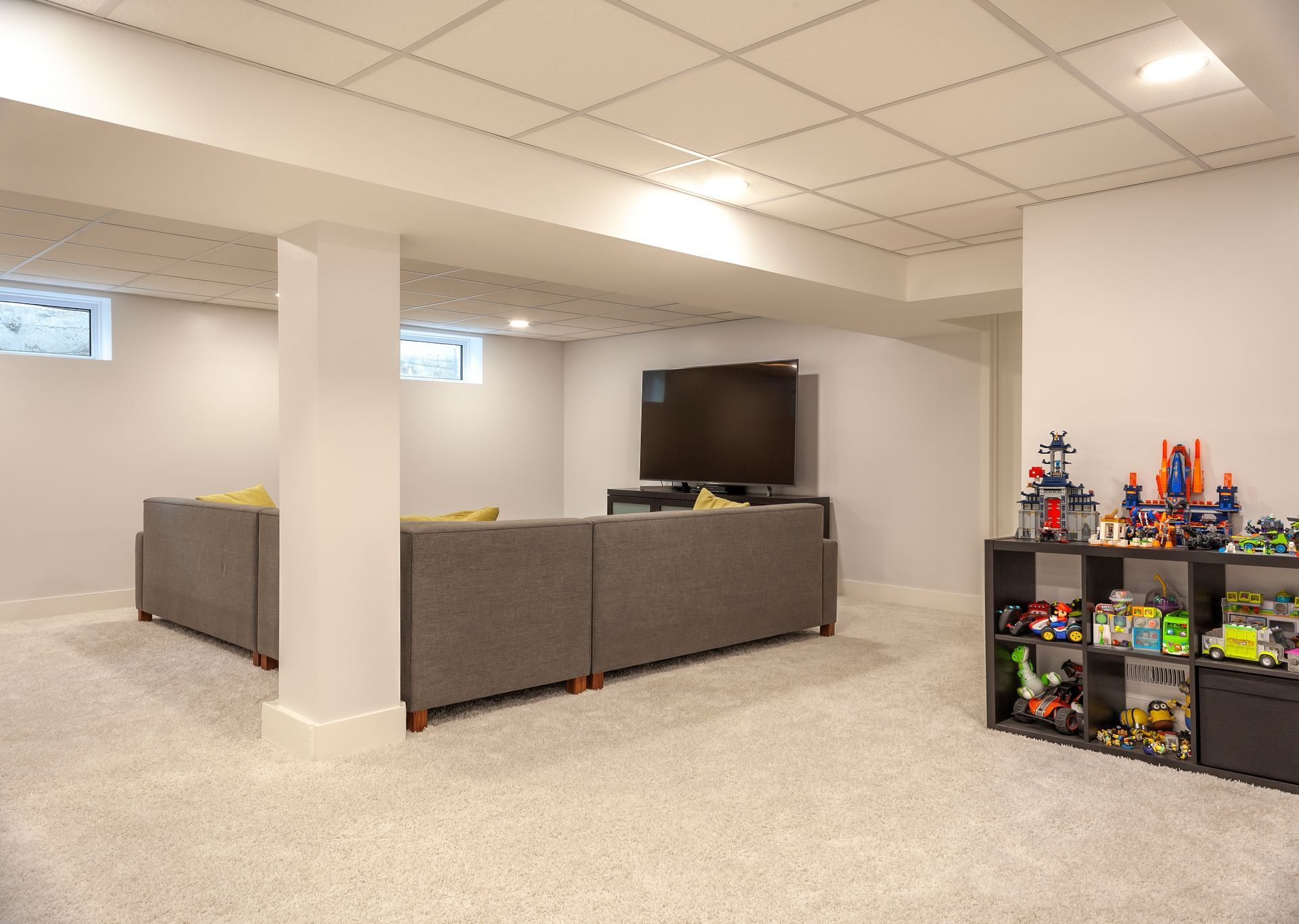 A finished basement with gray sectionals, a television on a stand, and a black storage shelf filled with colorful toys.